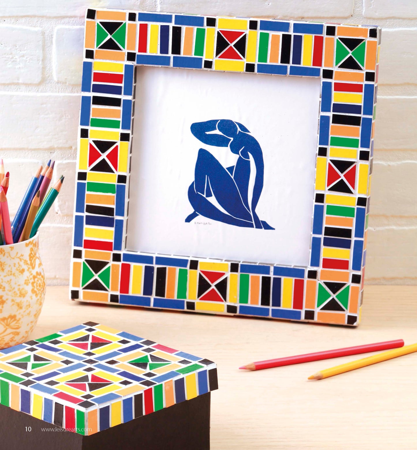 A colorful mosaic frame displays a blue abstract figure, showcasing creative patterns inspired by the Beginner’s Guide to Faux Mosaics from Leisure Arts. Next to it, colored pencils in a patterned cup and a coordinating mosaic box rest on a light wood surface—ideal inspiration for unique home decor projects.