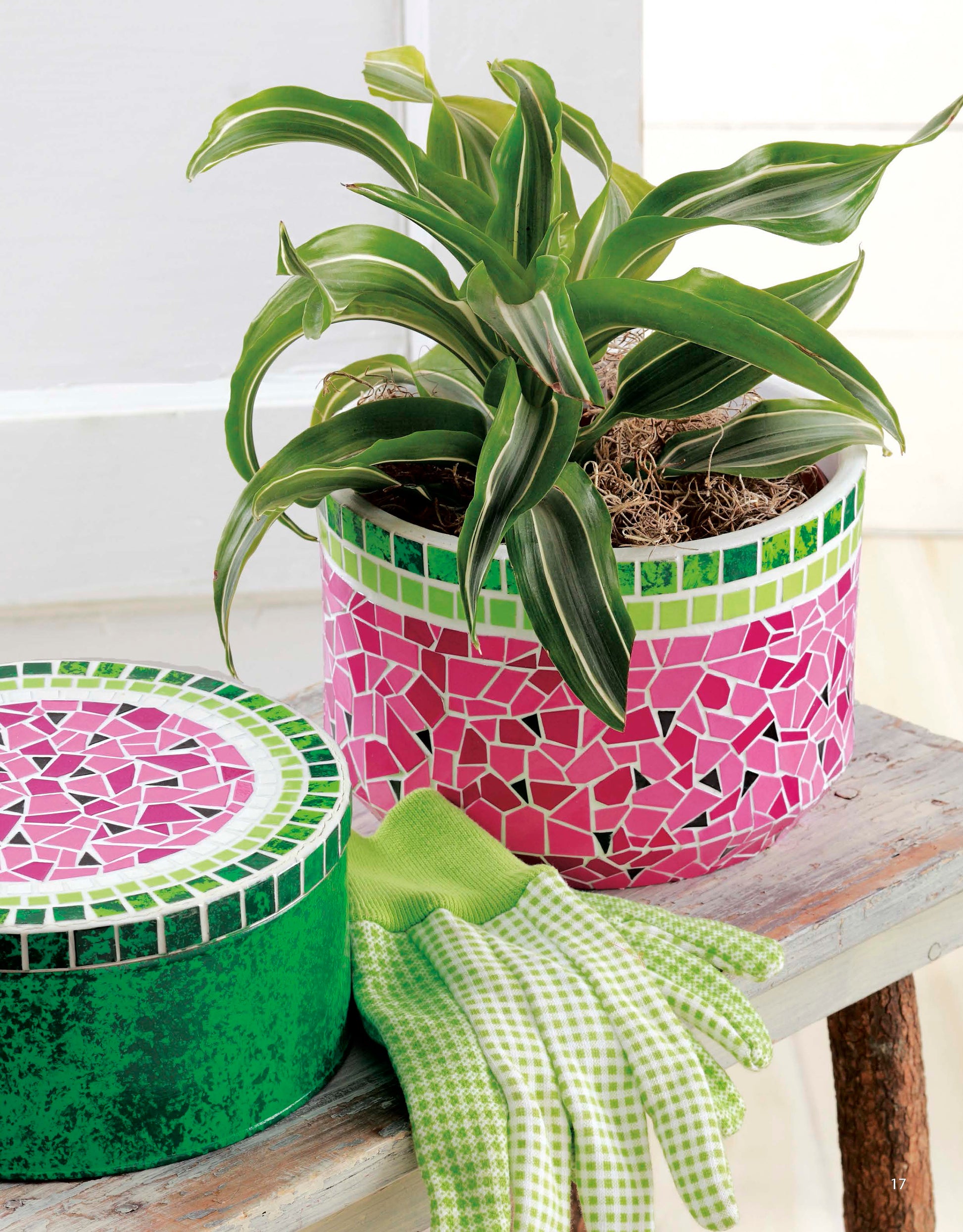 A copy of the “Beginner’s Guide to Faux Mosaics” by Leisure Arts rests on a wooden bench beside green and white gardening gloves and a green container with a mosaic lid, bringing home decor inspiration through intricate craft patterns.