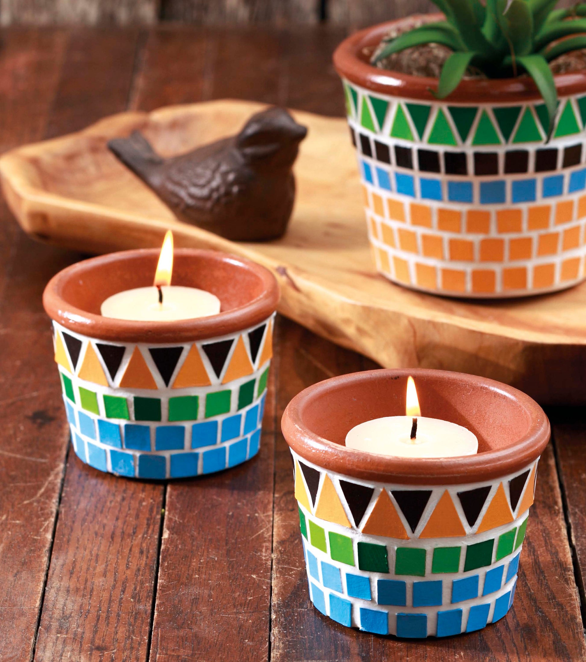 Two lit tealight candles in colorful mosaic-patterned holders rest on a wooden table, accompanied by a matching plant pot and small green plant, with a bird figurine in the background—an inviting scene that beautifully highlights unique home decor crafted using patterns from Leisure Arts’ Beginner’s Guide to Faux Mosaics.