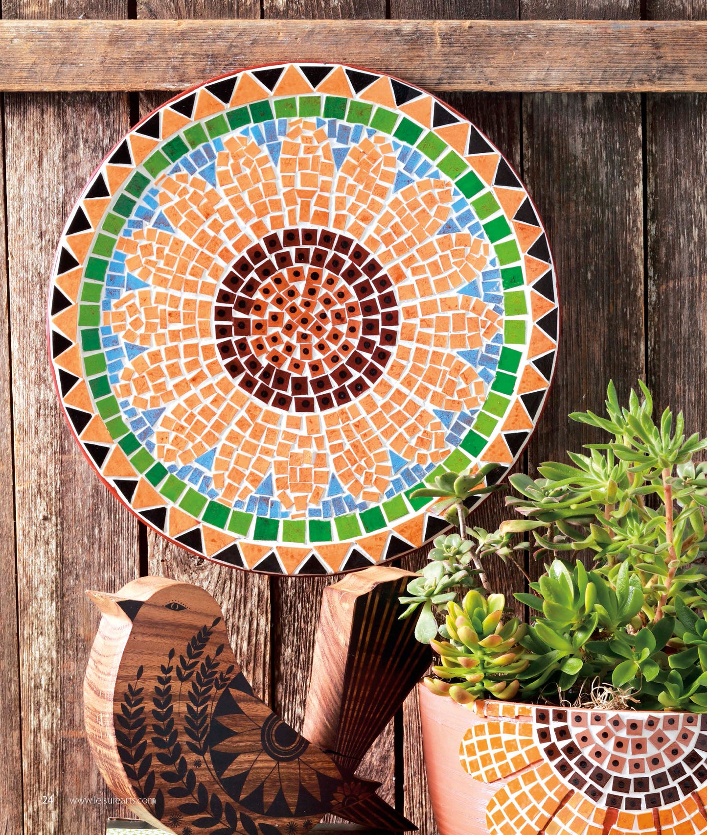 A vibrant sunflower mosaic plate, crafted using free-form techniques from Leisure Arts’ “Beginner’s Guide to Faux Mosaics,” decorates a wooden wall above a potted succulent and a small wooden bird figurine.