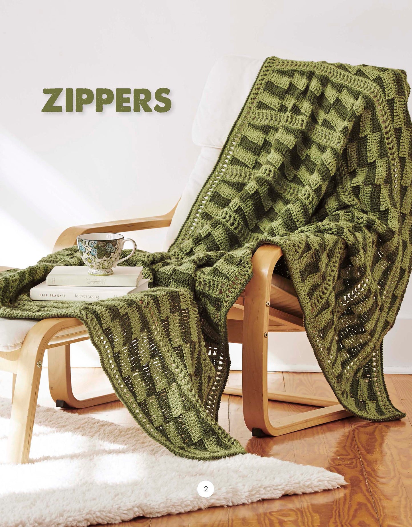 A green geometric-knit blanket from Leisure Arts' "Graphic Pattern Afghans" rests on a wooden chair with a white cushion, accompanied by a teacup and books—an inspiring scene for home decor projects.