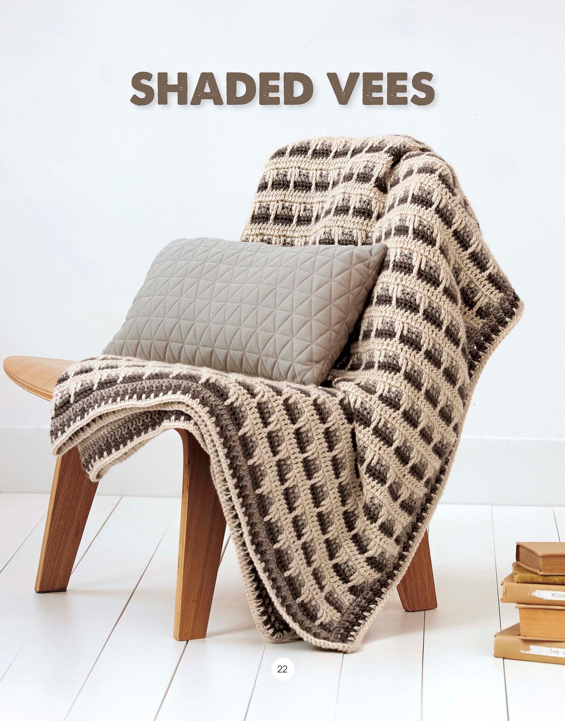 A modern wooden chair draped with a beige and brown crochet blanket from the Leisure Arts Graphic Pattern Afghans collection and a gray geometric pillow sits beside a stack of books—perfect inspiration for home decor. “SHADED VEES” appears above in bold letters.