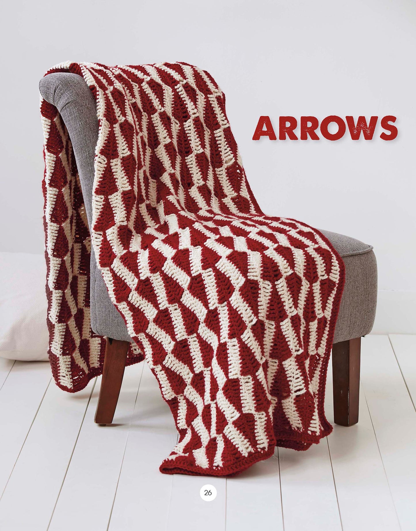 A Graphic Pattern Afghans blanket from Leisure Arts, featuring bold geometric designs in red and white, is draped over a gray chair. "ARROWS" in red letters adds style to this vibrant room with wood floors and chic home decor.