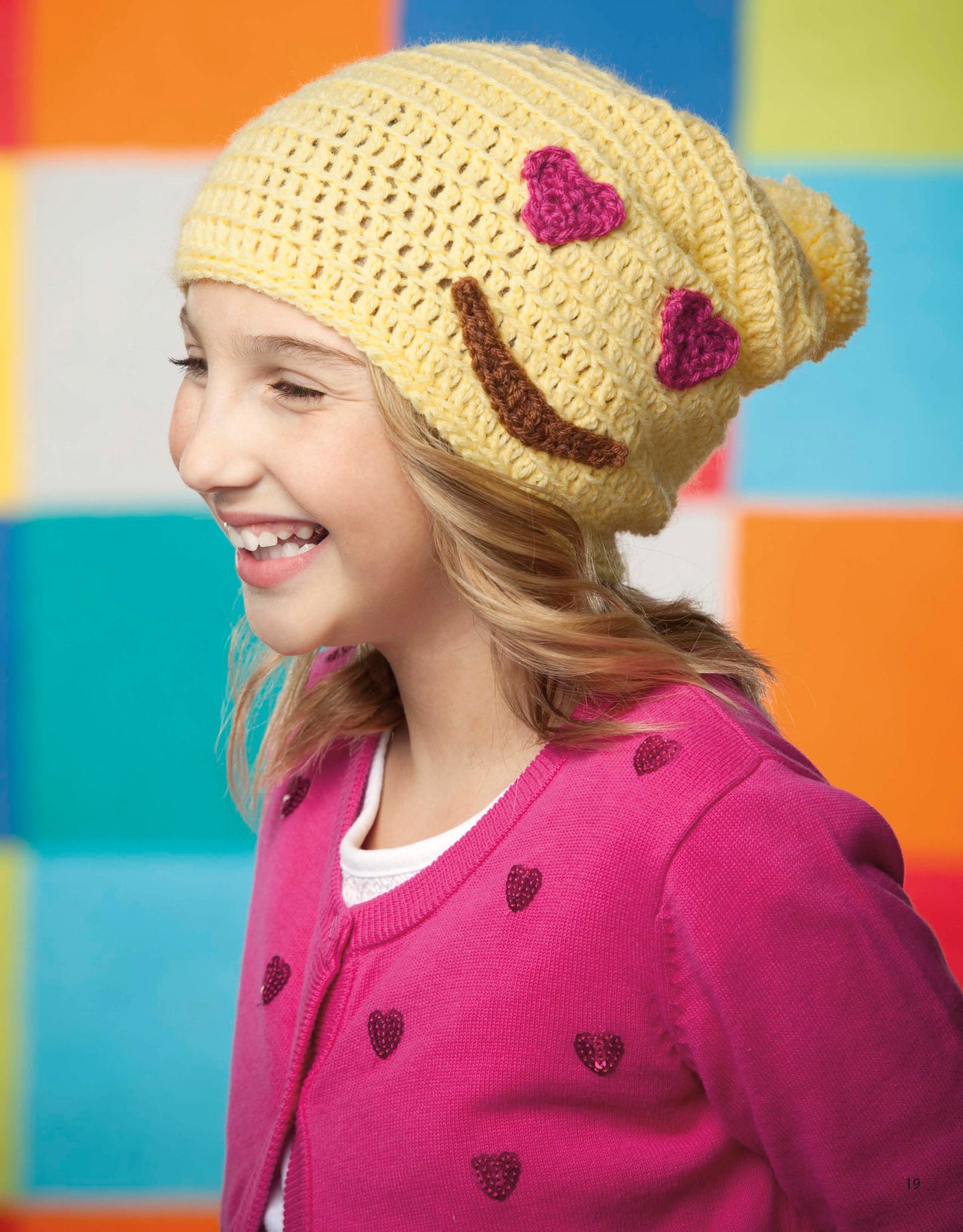 A smiling girl in a yellow beanie with a heart-eyes emoji and pink heart-pattern sweater stands before a colorful backdrop—perfect inspiration from Leisure Arts' "Emoji Crochet" for beginner crocheters.