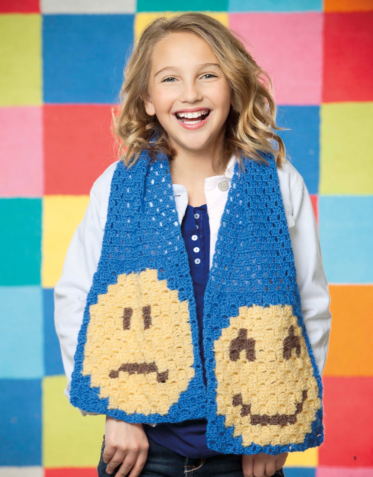 A smiling child models the blue "Emoji Crochet" scarf from Leisure Arts, featuring large happy and sad yellow faces, against a vibrant patchwork backdrop—a playful beginner project inspired by Leisure Arts patterns.