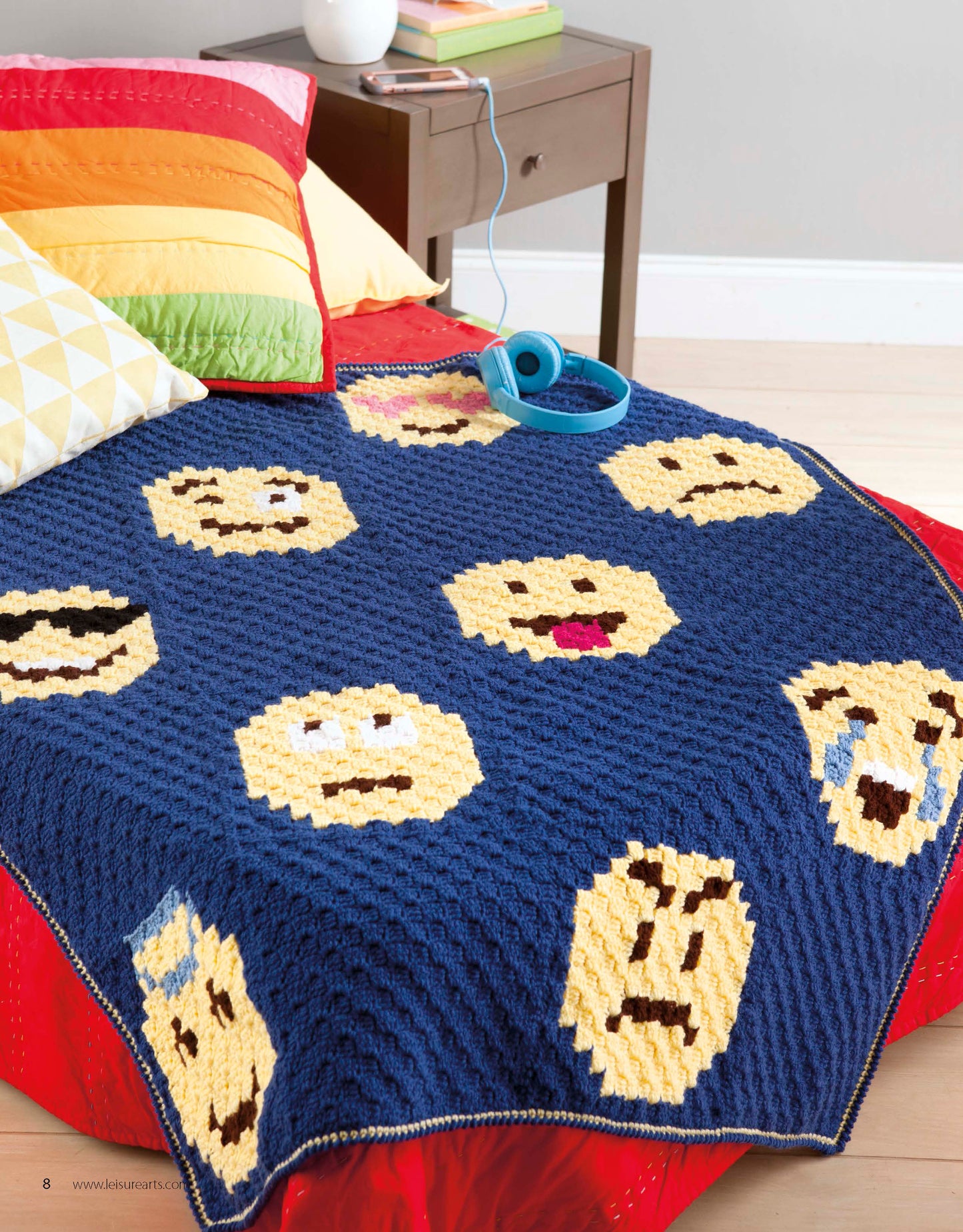 Leisure Arts’ Emoji Crochet blanket, featuring blue yarn and yellow emoji faces, is draped over a red bed with vibrant pillows. A blue headset rests nearby on a brown bedside table holding books and glasses—ideal for beginner projects.