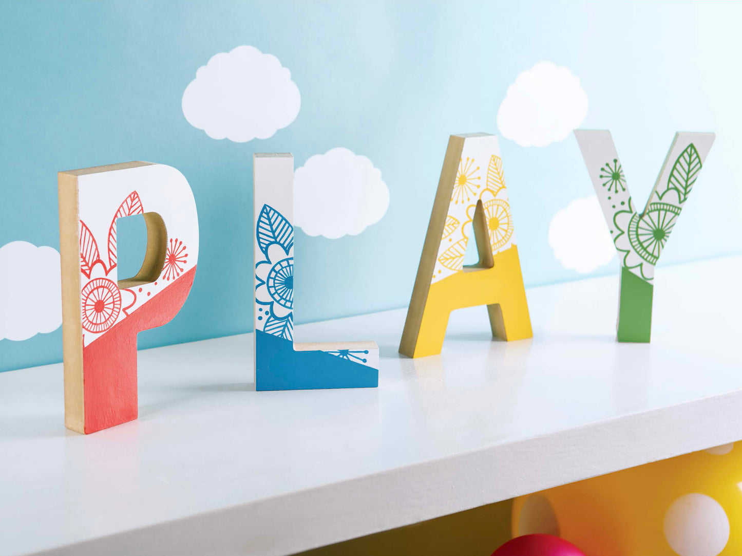 Brightly painted wooden letters spell out PLAY, each adorned with playful floral motifs and set on a white surface. Featured in Leisure Arts’ Initial It!—15 Projects, these eye-catching wood pieces are perfect for embellishing letters and sparking creativity in various crafts, from wreaths to pallet signs. A cheerful light blue background dotted with fluffy white clouds adds an inviting atmosphere to inspire your next project.