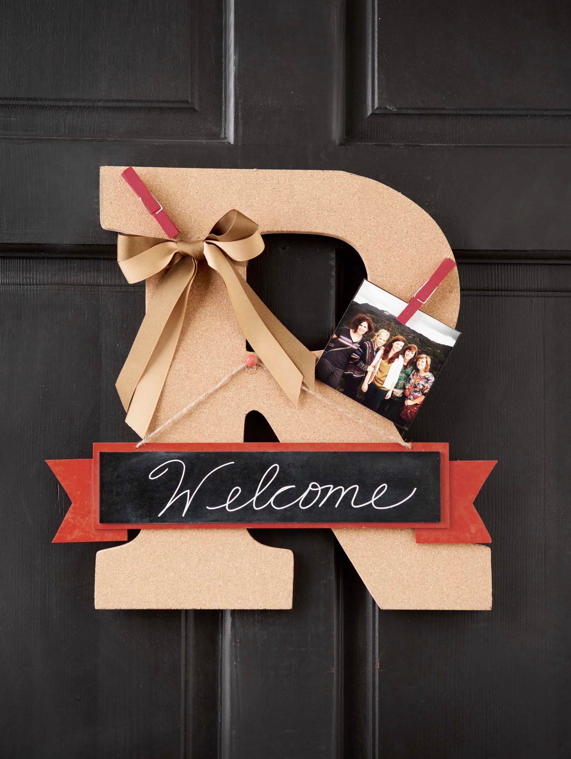 Personalize your decor by embellishing wood letters—like this large letter R cork board on a door, styled with a bow, Welcome chalkboard sign, and clipped family photo—for inspiration featured in Leisure Arts’ “Initial It!—15 Projects Using Simple Techniques to Embellish Wood Letters, Wreaths, Pallet Signs, and Other Crafty Accents.”.