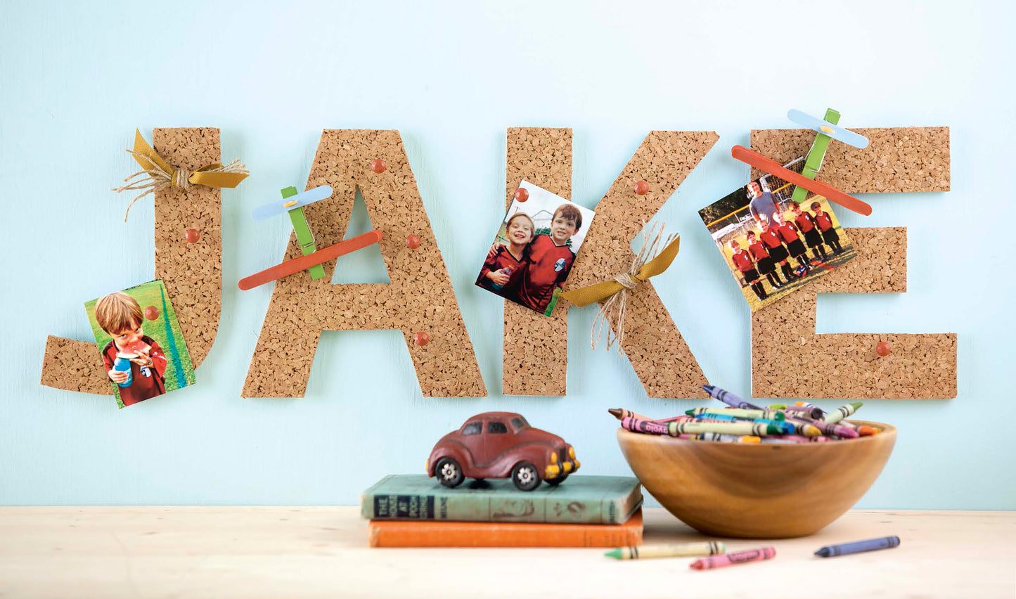 Discover creative inspiration with Leisure Arts’ Initial It!—15 Projects, a guide featuring simple techniques to embellish wood letters, wreaths, pallet signs, and more. Here, “JAKE” is spelled in cork letters decorated with photos, popsicle sticks, and wheat—just one example of the unique projects you can try. Displayed in front are crayons, a toy car on books, and a wooden bowl of crayons for added crafty flair.