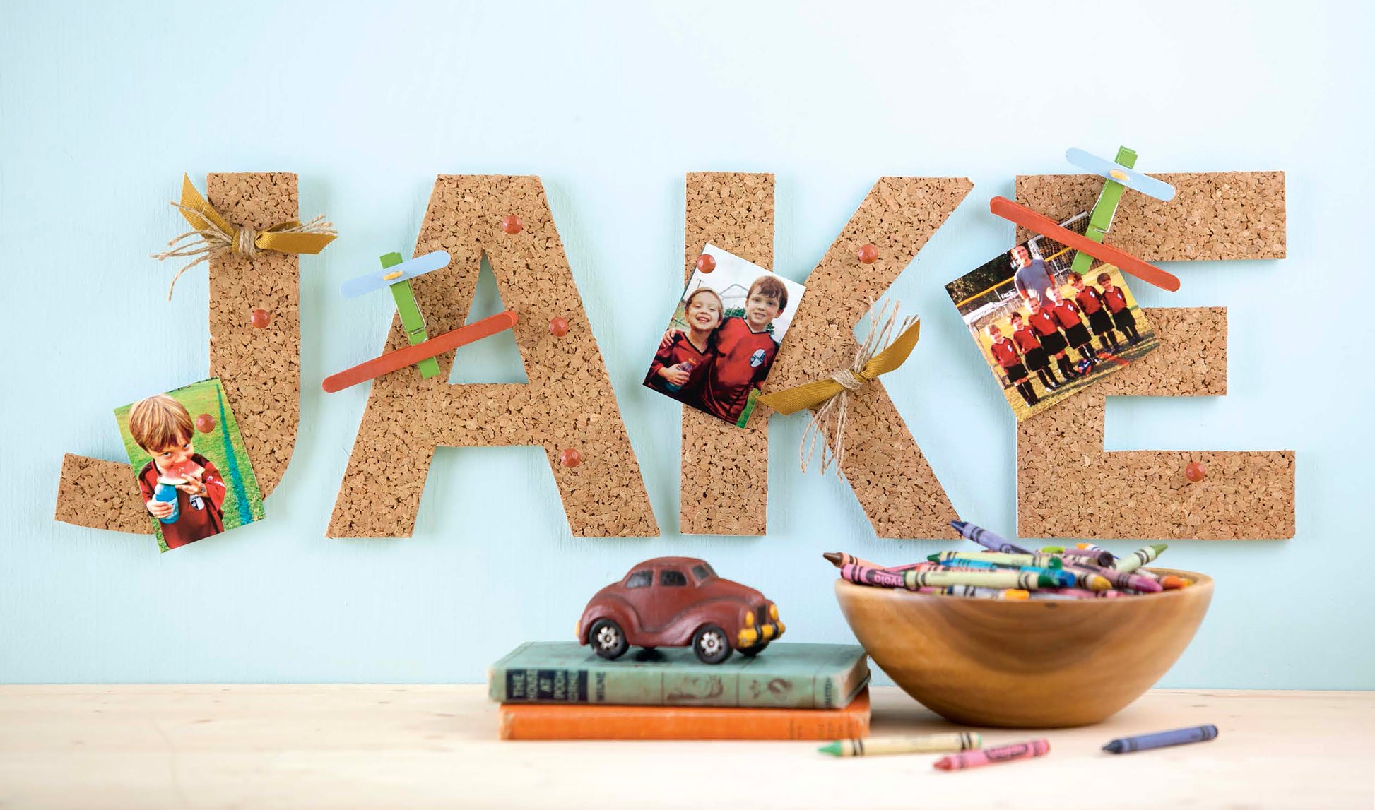 Discover creative inspiration with Leisure Arts’ Initial It!—15 Projects, a guide featuring simple techniques to embellish wood letters, wreaths, pallet signs, and more. Here, “JAKE” is spelled in cork letters decorated with photos, popsicle sticks, and wheat—just one example of the unique projects you can try. Displayed in front are crayons, a toy car on books, and a wooden bowl of crayons for added crafty flair.