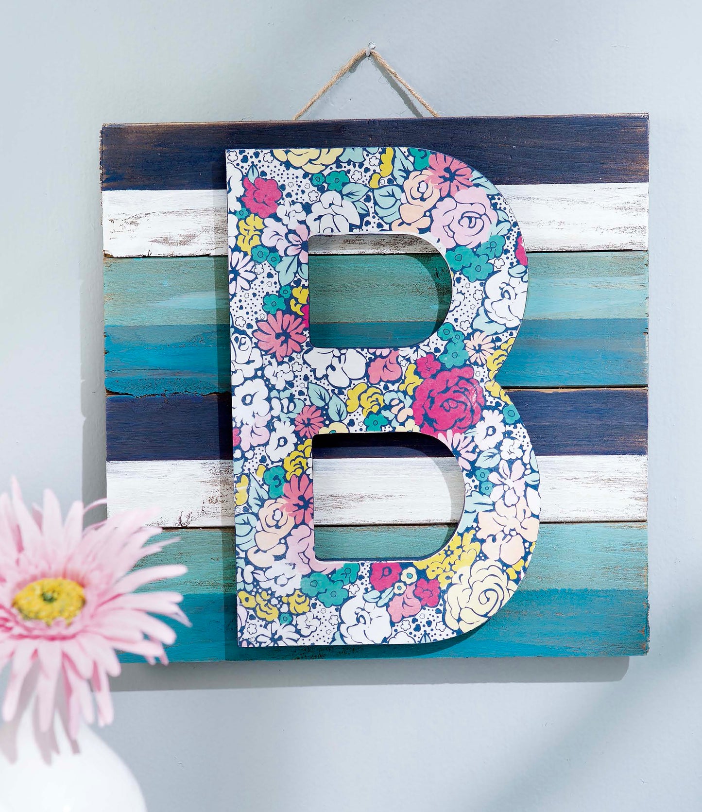 Discover creative ways to embellish wood letters, wreaths, pallet signs, and more with “Initial It!” by Leisure Arts. One of the 15 simple projects features a wooden pallet sign decorated with horizontal blue, white, and teal stripes and an eye-catching floral-patterned letter B. Add vibrant prints and accents to your crafts—a pretty pink flower in the bottom left corner offers a charming finishing touch to any project!.