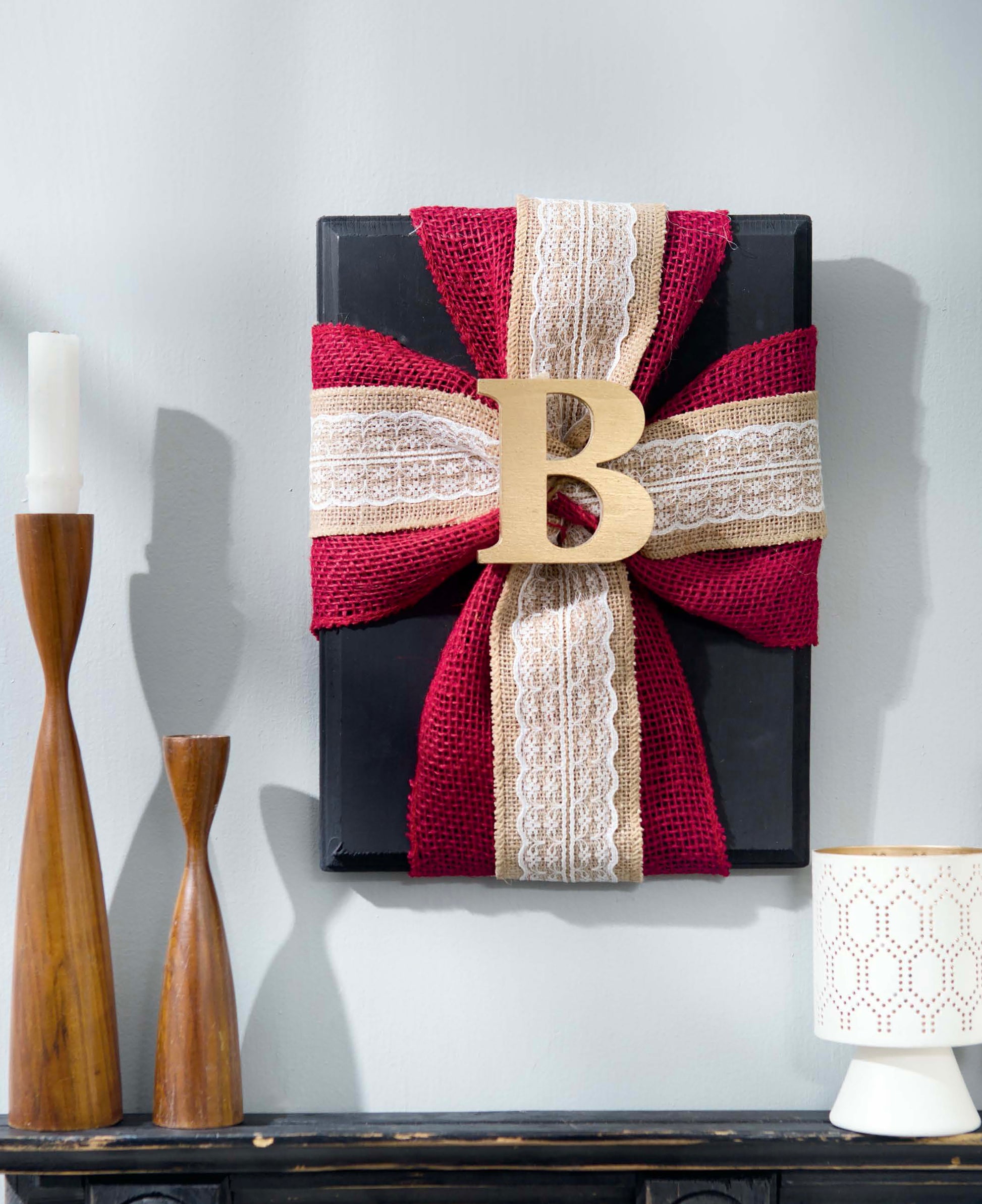 A black rectangular gift with a large bow crafted from red, lace, and burlap ribbons is beautifully accented with a wooden letter B—an inspiring project idea from Leisure Arts’ “Initial It!—15 Projects,” which showcases creative ways to embellish wood letters for charming crafts. Nearby, wooden candlesticks and a white decorative candle holder complete the stylish arrangement.