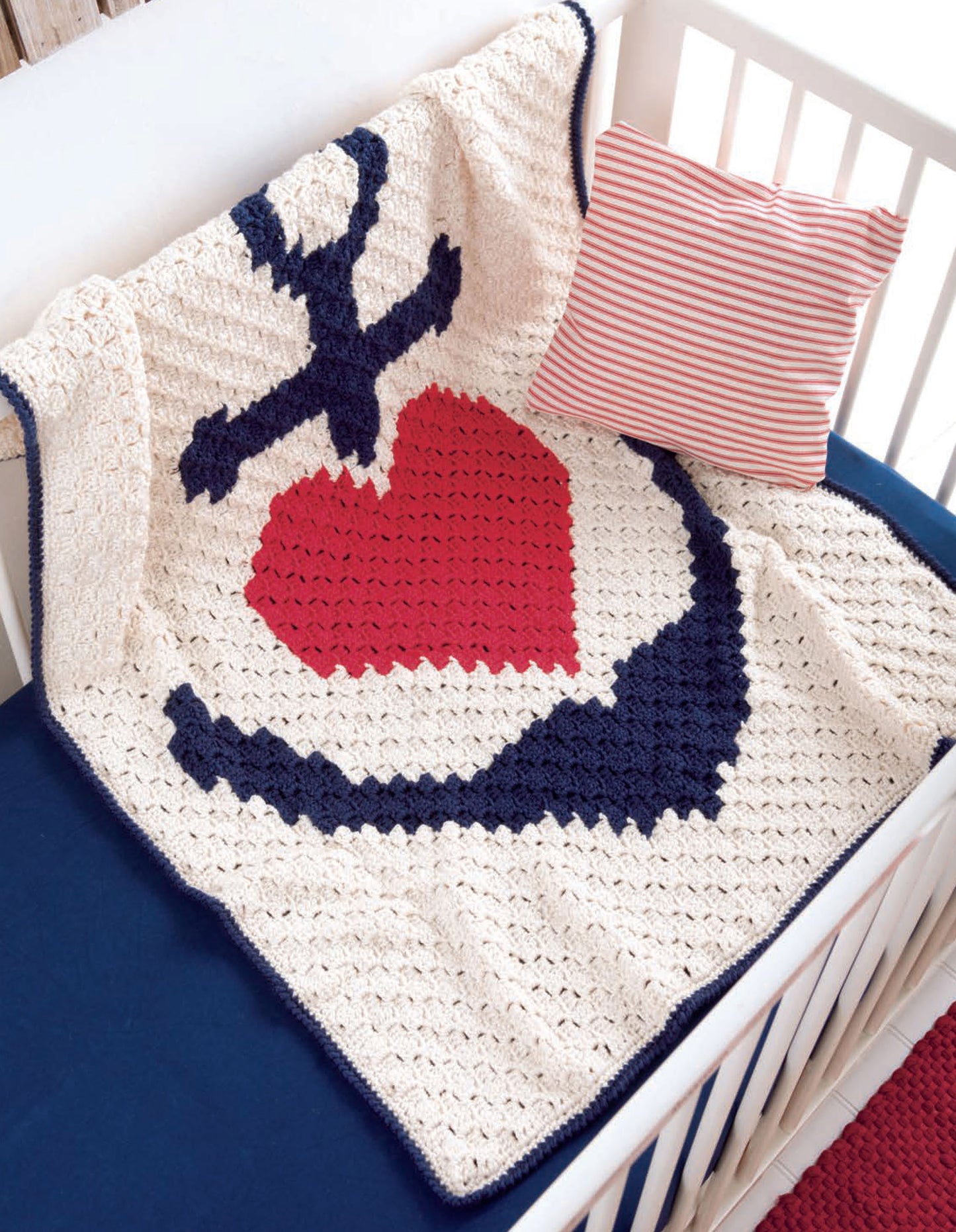 The Corner to Corner Baby Afghans by Leisure Arts features a navy anchor and red heart design draped over a white crib with solid navy bedding and a red-and-white striped pillow—ideal for fans of easy crochet patterns.