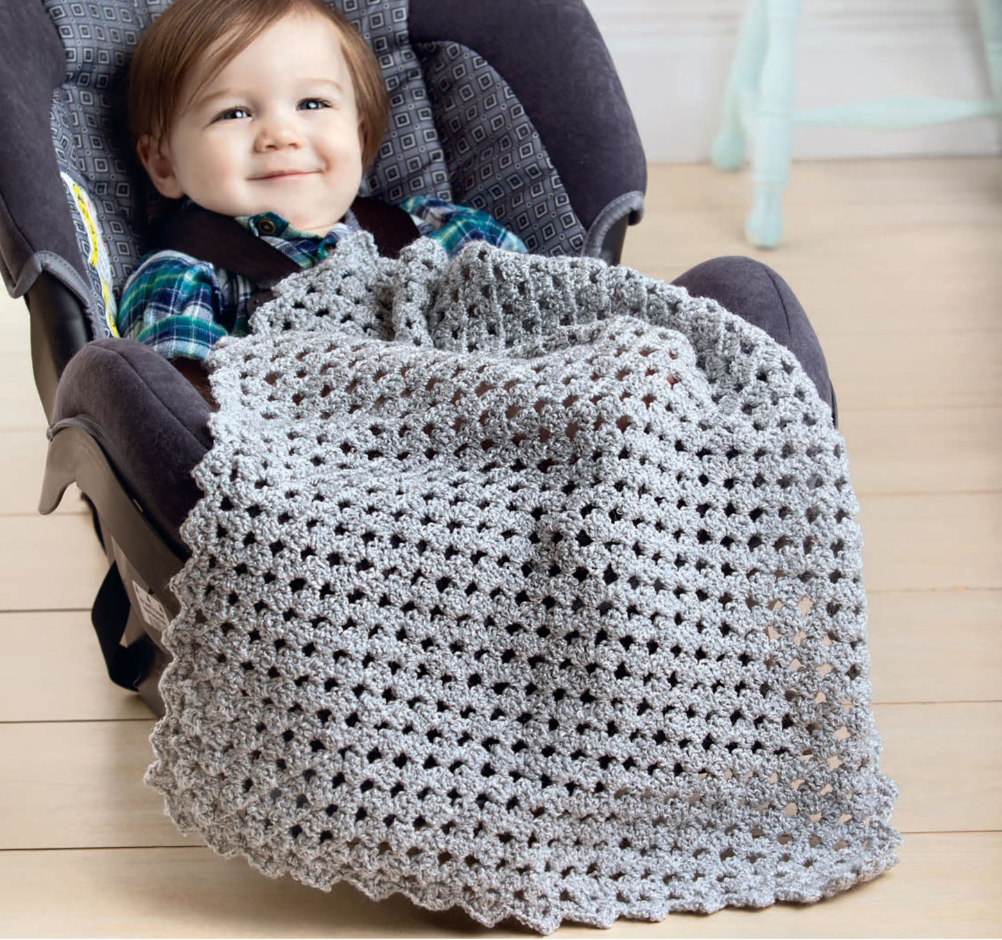 A smiling baby sits in a car seat, covered with a light gray afghan made from Leisure Arts' Corner to Corner Baby Afghans. The car seat has a patterned lining, and the scene is indoors on a wooden floor.