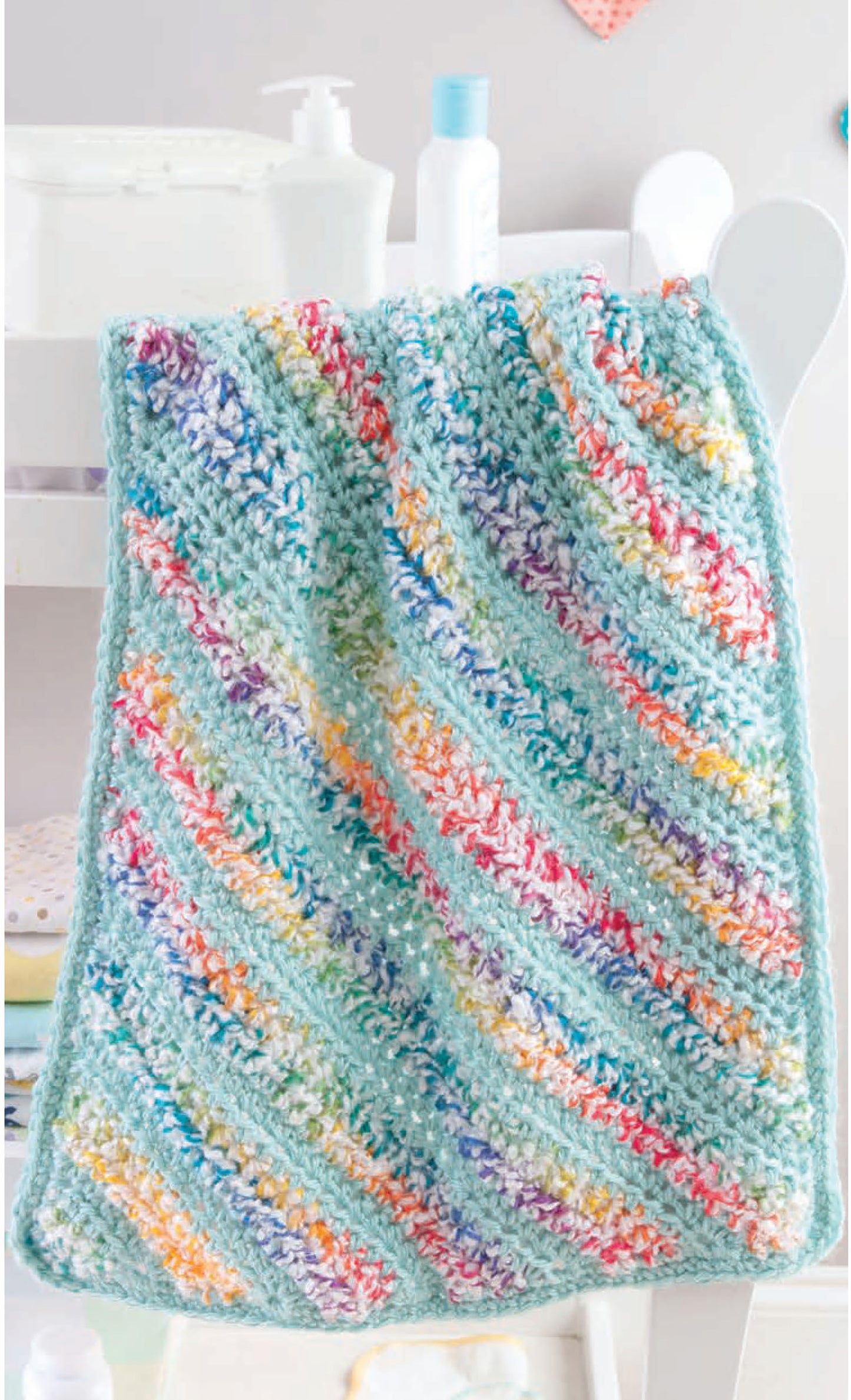 The Leisure Arts Corner to Corner Baby Afghans showcases a soft crocheted blanket with diagonal pastel stripes in blue, yellow, orange, purple, and green, draped over a white chair with baby items blurred in the background.