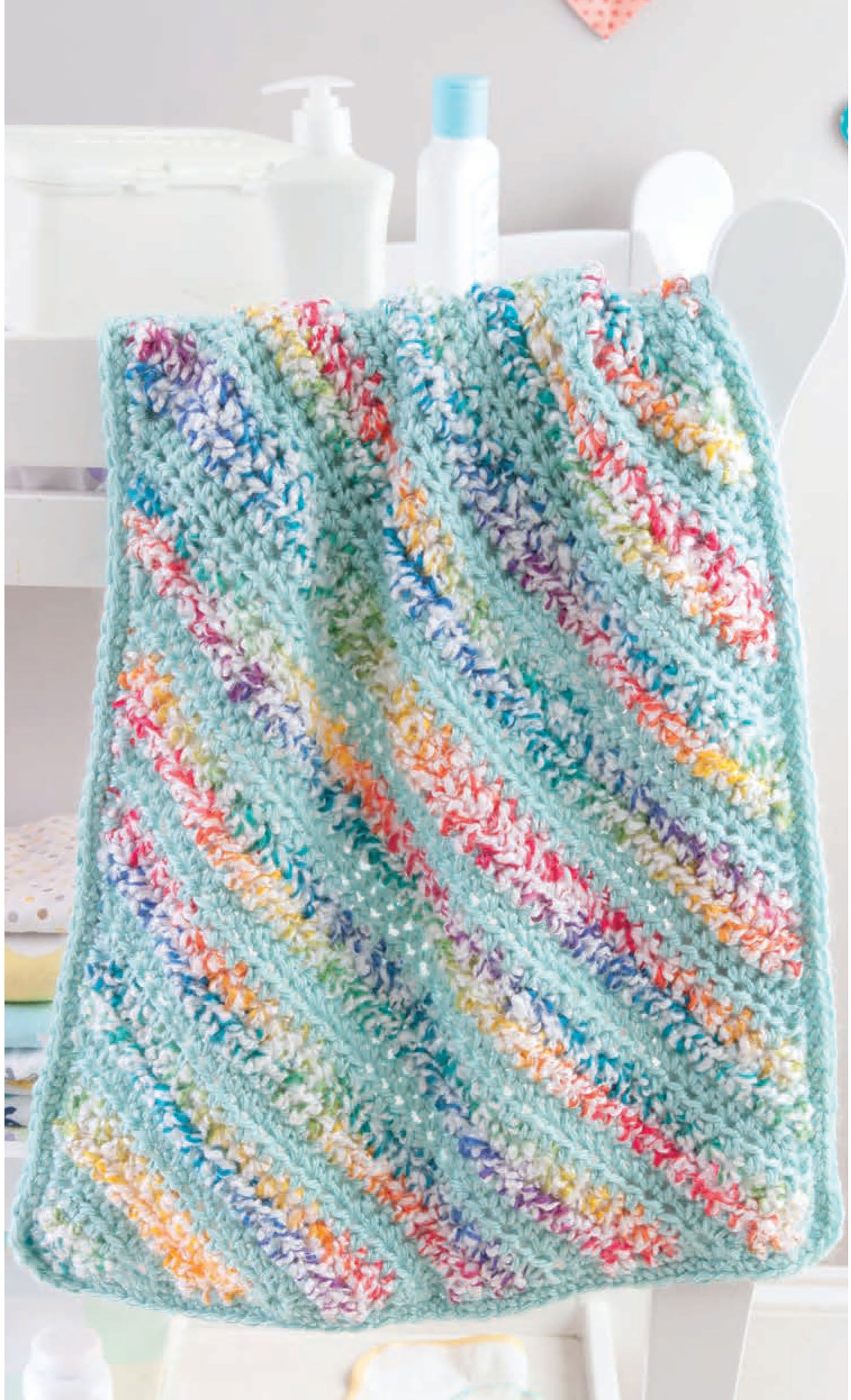 The Leisure Arts Corner to Corner Baby Afghans showcases a soft crocheted blanket with diagonal pastel stripes in blue, yellow, orange, purple, and green, draped over a white chair with baby items blurred in the background.