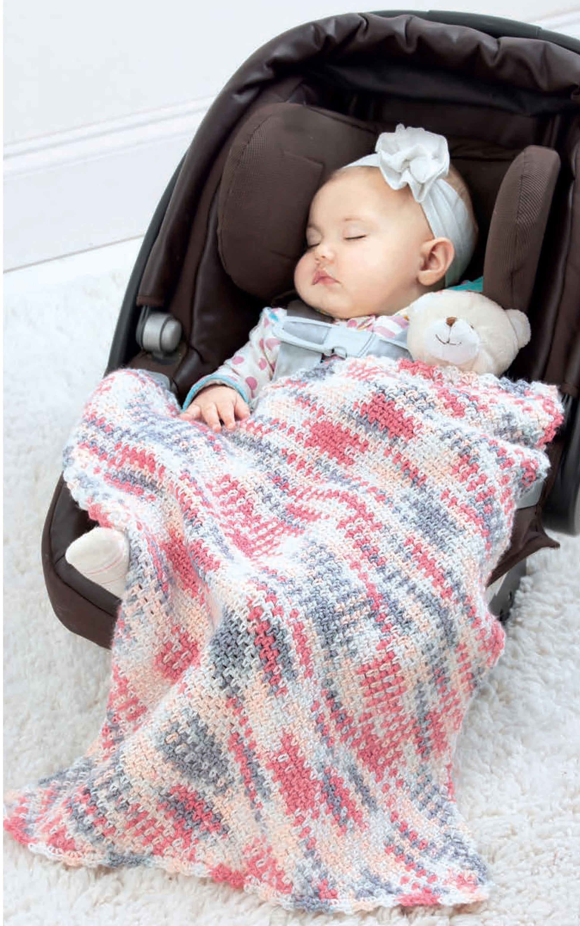 A baby sleeps in a car seat under a pink, gray, and white Leisure Arts Corner to Corner Baby Afghan, holding a stuffed bear and wearing a bow headband.