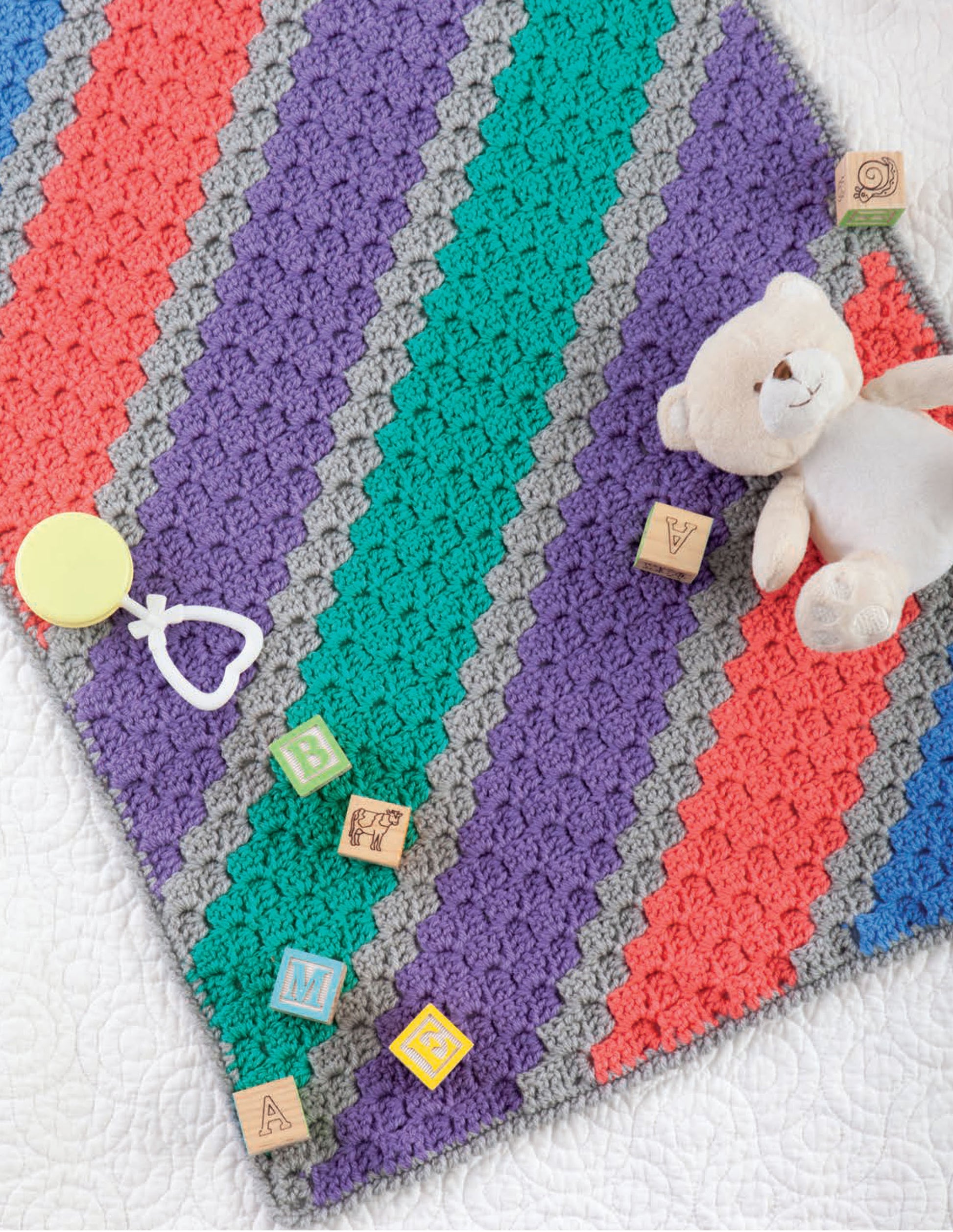 The Corner to Corner Baby Afghans by Leisure Arts features a crocheted blanket with red, teal, purple, and gray stripes on a white surface. Wooden alphabet blocks, a pale yellow pacifier, and a beige teddy bear rest on top—ideal for crochet enthusiasts.