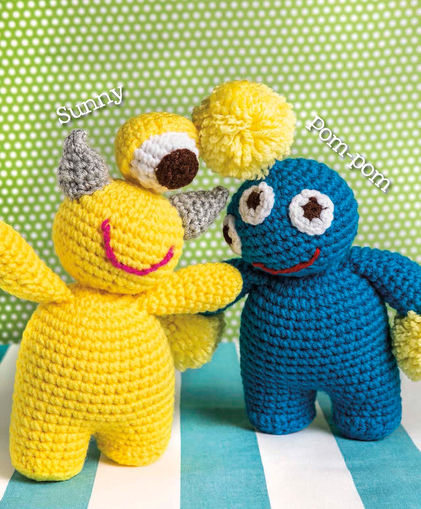 Two crocheted plush toys from Leisure Arts’ Cute Amigurumi Monsters Digital Download stand side by side: a yellow, one-eyed monster with gray horns labeled “Sunny,” and a blue, two-eyed monster with a yellow pom-pom head labeled “pom-pom.”.