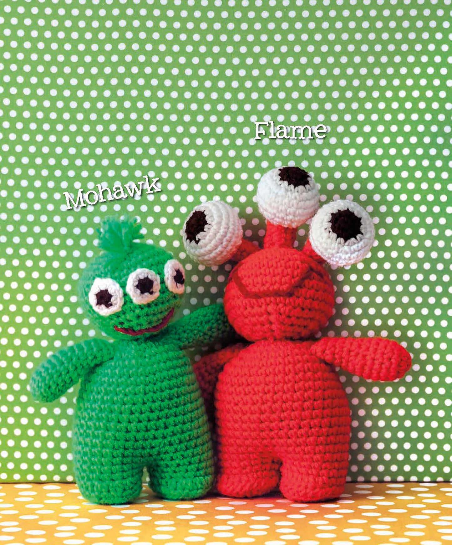 Two cute amigurumi monsters from Leisure Arts’ "Cute Amigurumi Monsters Digital Download" stand together, Mohawk is green with three eyes and a hair tuft, while Flame is red with three quirky eye stalks.