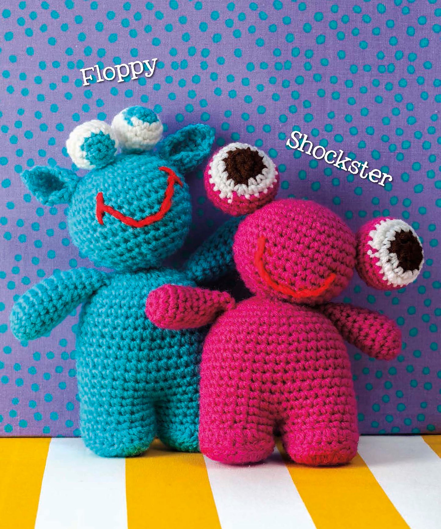 Two amigurumi dolls from Leisure Arts' Cute Amigurumi Monsters Digital Download stand side by side—one blue with three eyes ("Floppy") and one pink with two big eyes ("Shockster")—on a purple, blue polka-dotted background.