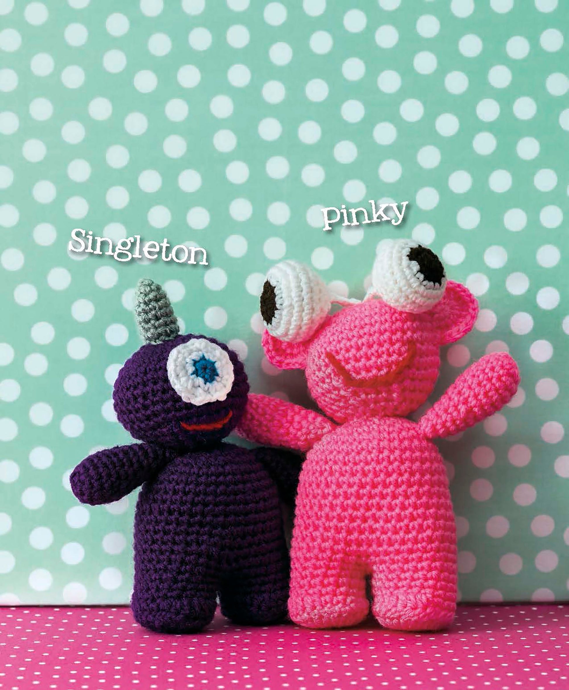 Singleton, a purple amigurumi with one blue eye and gray horn, and Pinky, a pink plush with big googly eyes, sit together in front of a turquoise polka dot background. Pattern: Cute Amigurumi Monsters Digital Download by Leisure Arts.