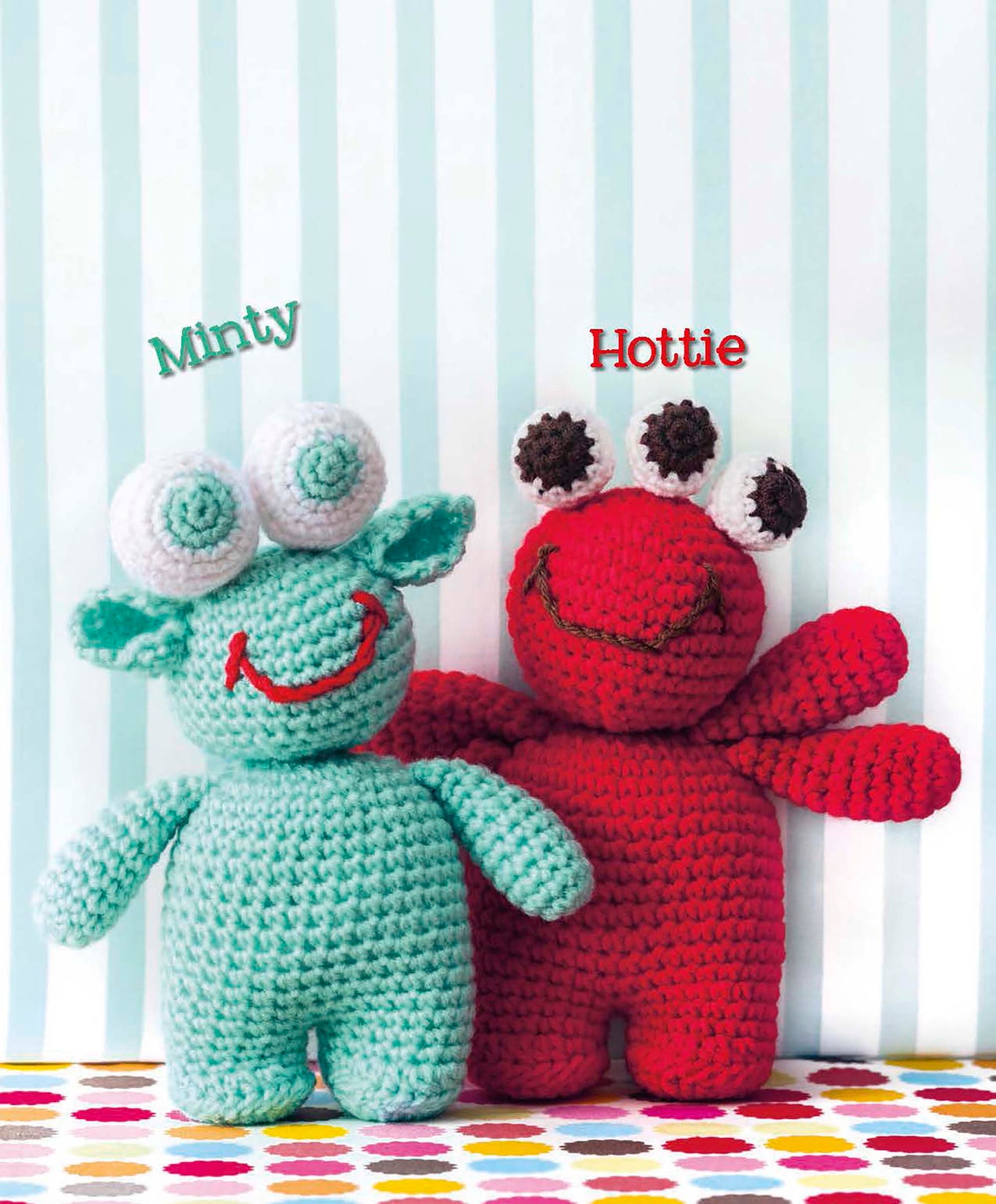 Two crocheted amigurumi monsters, “Minty” in aqua with three eyes and ears, and “Hottie” in red with wide arms, stand side by side. Download this Cute Amigurumi Monsters pattern by Leisure Arts to create your own unique plush toys!.