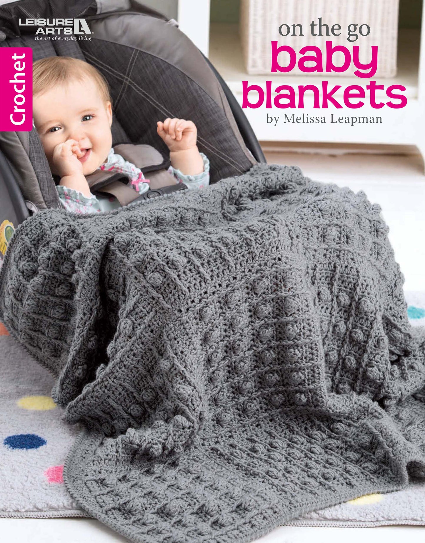 A smiling baby sits in a car seat, wrapped in a large, textured gray baby blanket featuring bobble and cable crochet. Text reads: On the Go Baby Blankets Digital Download by Leisure Arts.