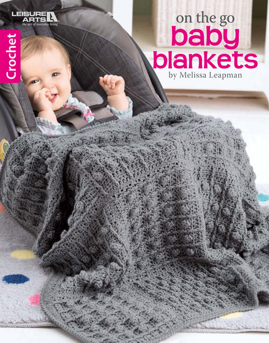 A smiling baby sits in a car seat, wrapped in a large, textured gray baby blanket featuring bobble and cable crochet. Text reads: On the Go Baby Blankets Digital Download by Leisure Arts.