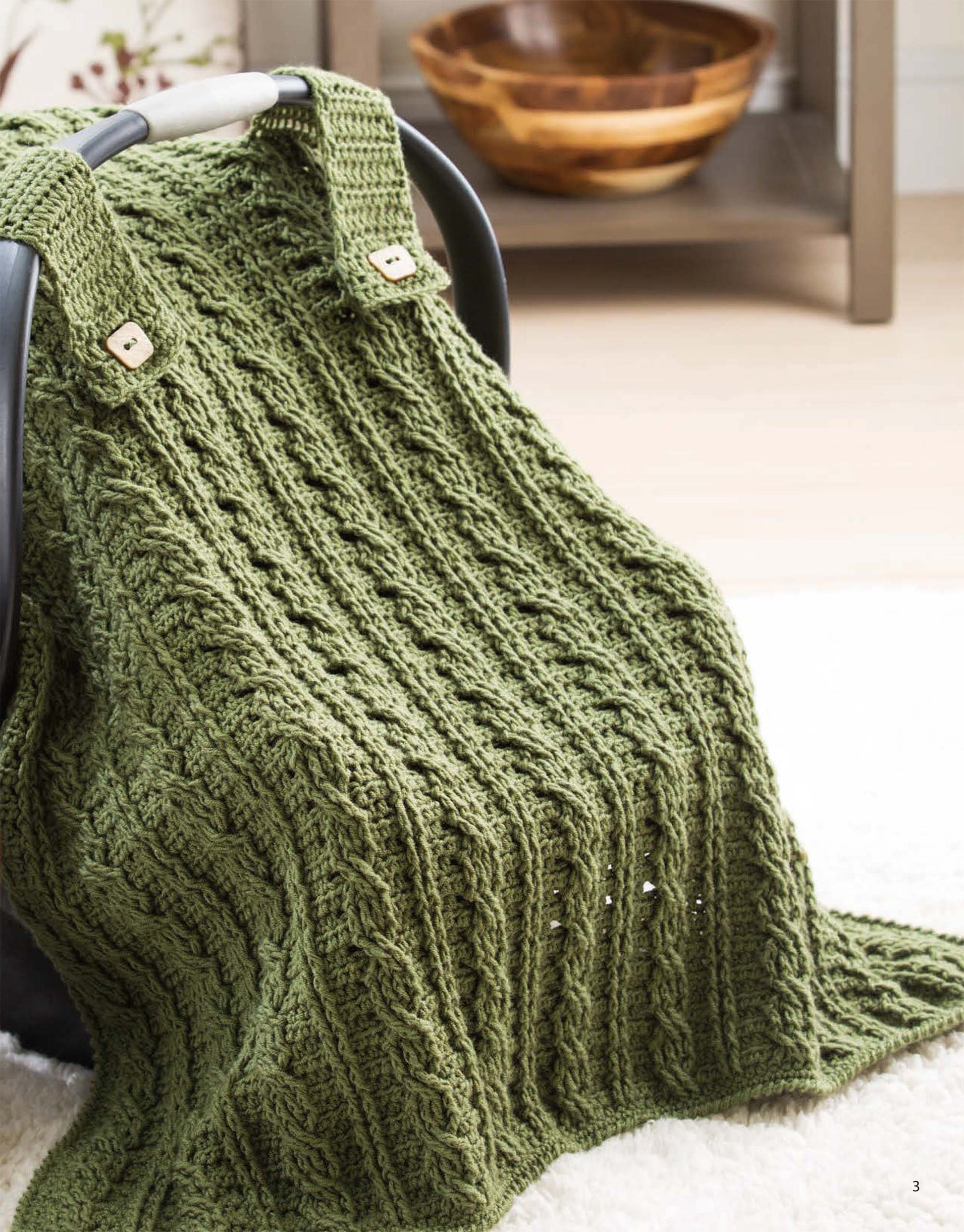 The On the Go Baby Blankets Digital Download by Leisure Arts features a green, textured knit car seat blanket with cable pattern and buttoned straps. A wooden bowl sits on a shelf in the blurred background for crochet inspiration.