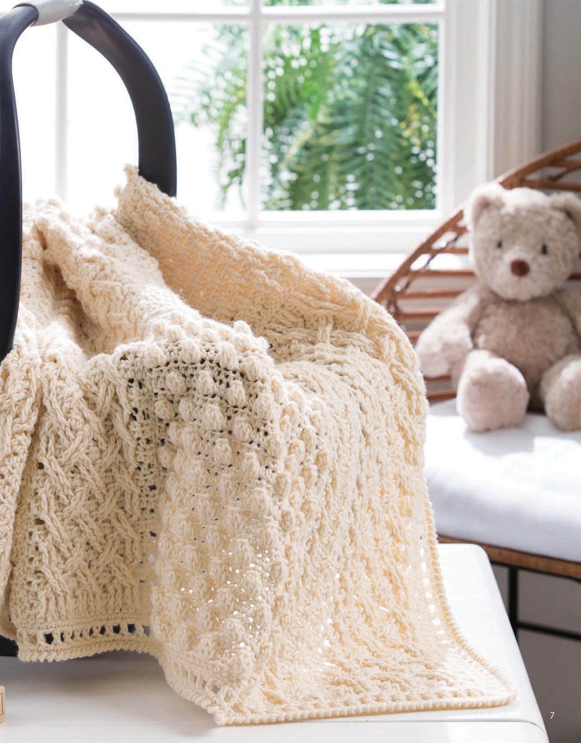 A cream-colored baby car seat blanket from Leisure Arts' "On the Go Baby Blankets" digital download drapes over the handle, with a plush teddy bear on a bench by a window overlooking lush green foliage.