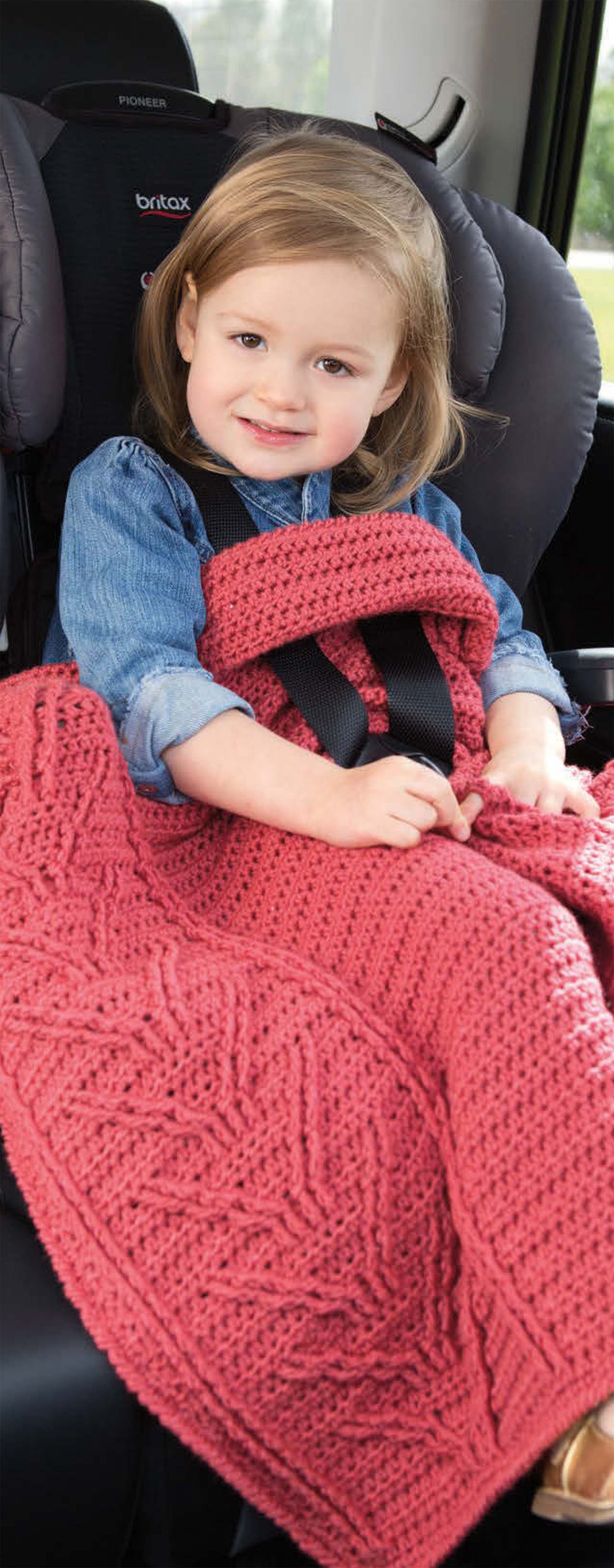 A young child smiles in her car seat, wearing a blue denim top and covered with the On the Go Baby Blankets Digital Download by Leisure Arts, inspired by crochet patterns. Her seatbelt fits securely over the cozy, textured blanket.