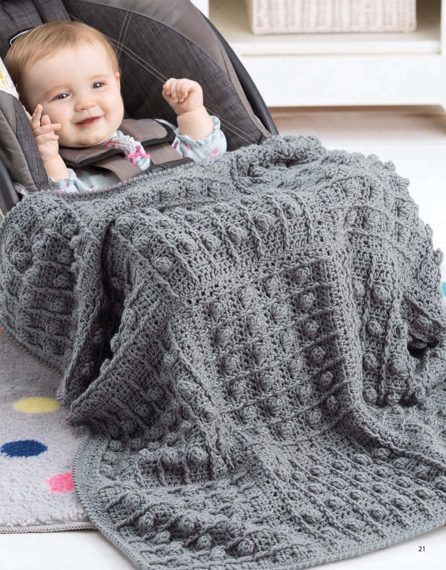 A smiling baby sits in a car seat, snug in the textured gray "On the Go Baby Blankets Digital Download" by Leisure Arts. The soft rug beneath, dotted with color, adds warmth and charm to the cozy scene.