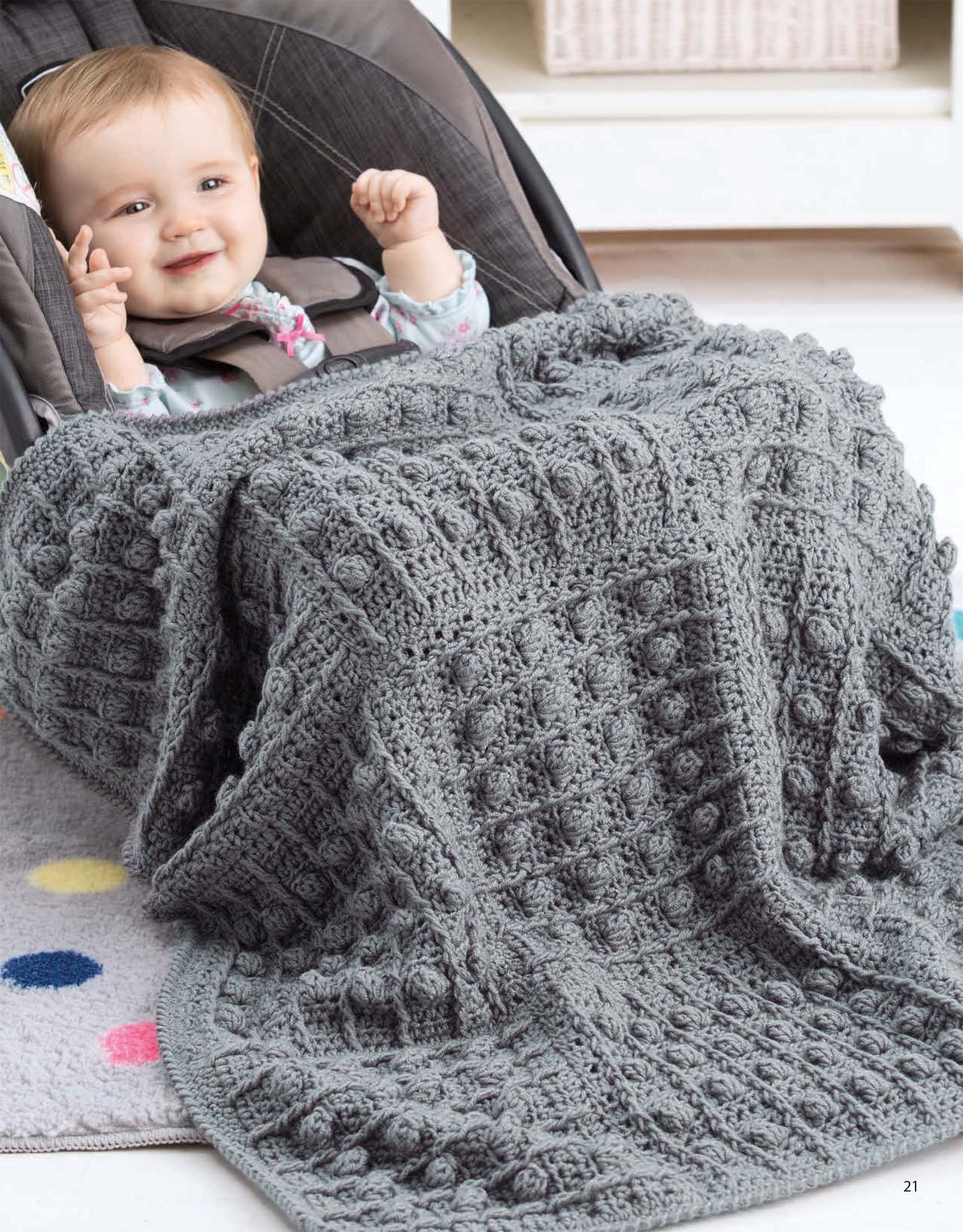 A smiling baby sits in a car seat, snug in the textured gray "On the Go Baby Blankets Digital Download" by Leisure Arts. The soft rug beneath, dotted with color, adds warmth and charm to the cozy scene.