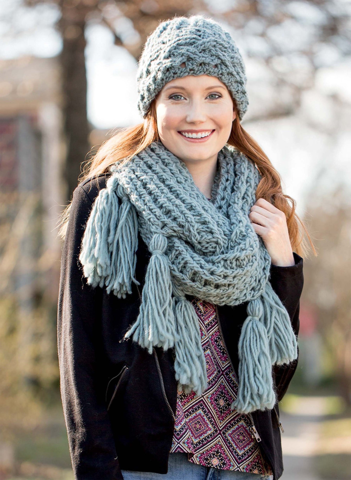 A smiling person outdoors wears a chunky blue knit hat and tassel scarf—just like the projects in the Textured Hats, Scarves, & Cowls Digital Download by Leisure Arts. Cozy inspiration in a black jacket with trees and a blurred building behind.
