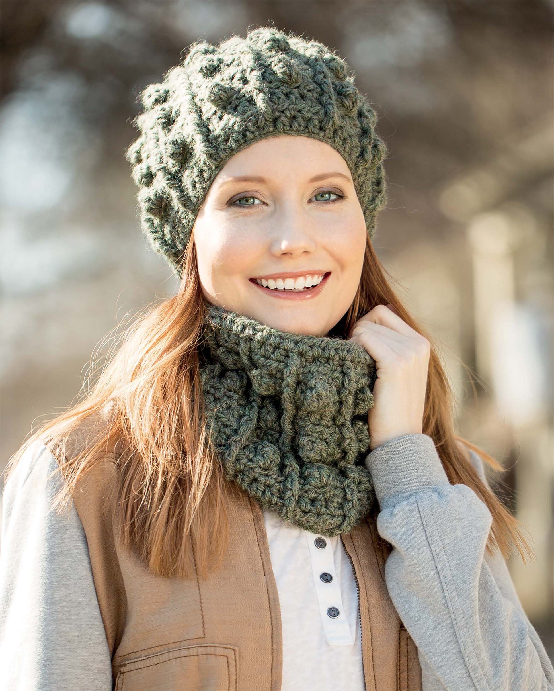 A smiling woman with long auburn hair models a textured green knit hat and matching cowl outdoors, featuring the "Textured Hats, Scarves, & Cowls Digital Download" by Leisure Arts. She wears a light brown vest over a gray hoodie against a blurred background.