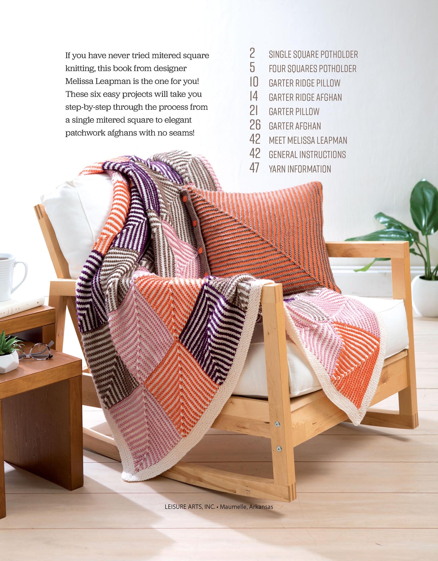 A wooden chair with a white cushion showcases the Leisure Arts Mitered Square Knitting Digital Download blanket and matching pillow in orange, pink, brown, and cream. Nearby, a table holds a plant and magazines; book contents are listed on the wall.