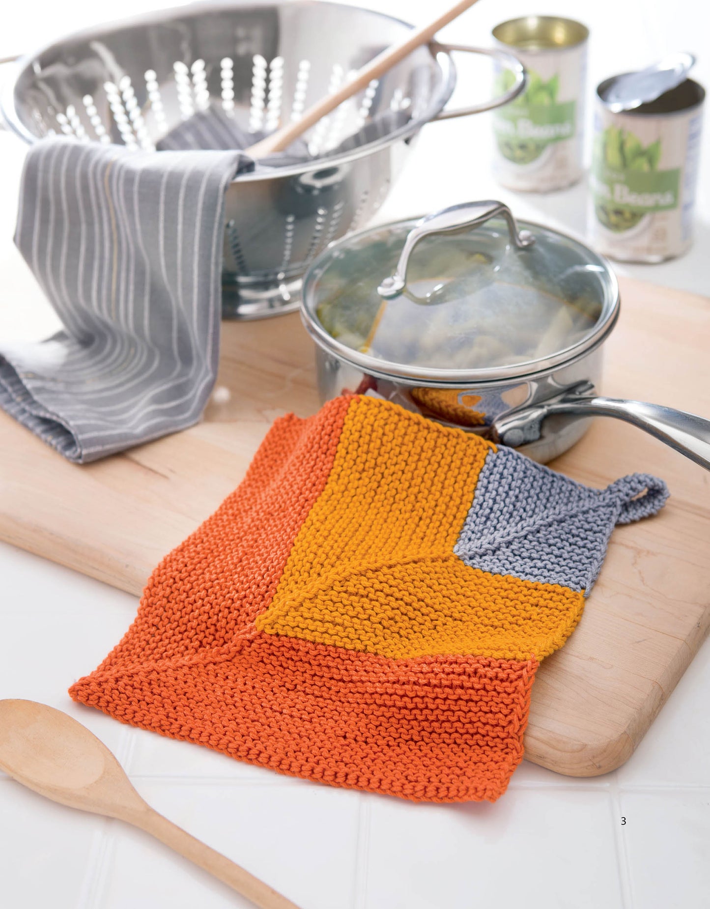 The Mitered Square Knitting Digital Download by Leisure Arts features a colorful orange, yellow, and gray garter-stitch pot holder displayed on a cutting board near a saucepan, striped towel, wooden spoon, colander, and canned goods.
