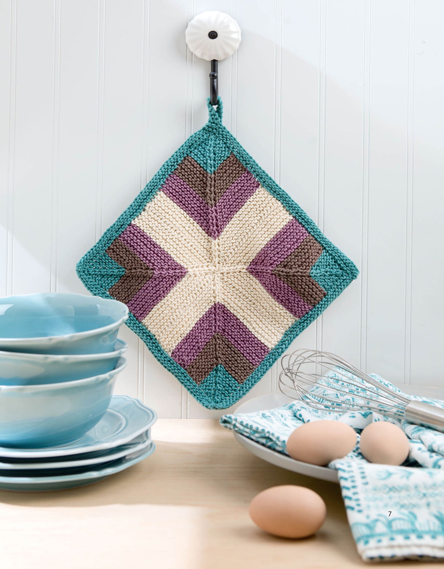 A colorful, diamond-shaped potholder made using the Mitered Square Knitting Digital Download by Leisure Arts hangs on a hook above a kitchen counter with blue dishes, eggs, and a whisk in a patterned bowl against a white paneled wall.