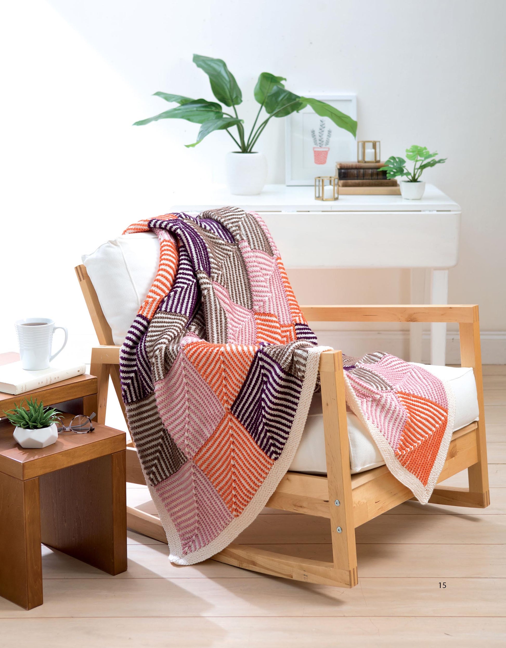 A cozy chair draped with the Mitered Square Knitting Digital Download by Leisure Arts sits next to a wooden table with a mug and books. A white desk, plants, and framed print complete the bright and inviting room.