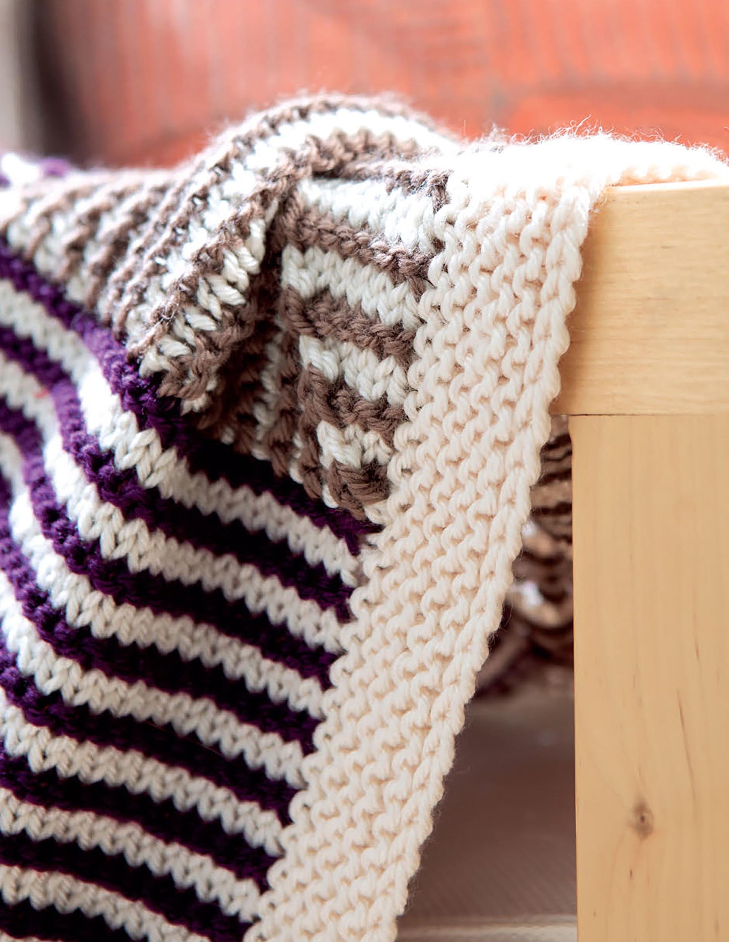 The Mitered Square Knitting Digital Download from Leisure Arts features a patchwork afghan with brown, white, and dark purple stripes plus a thick cream border, draped over the edge of a light wooden table.