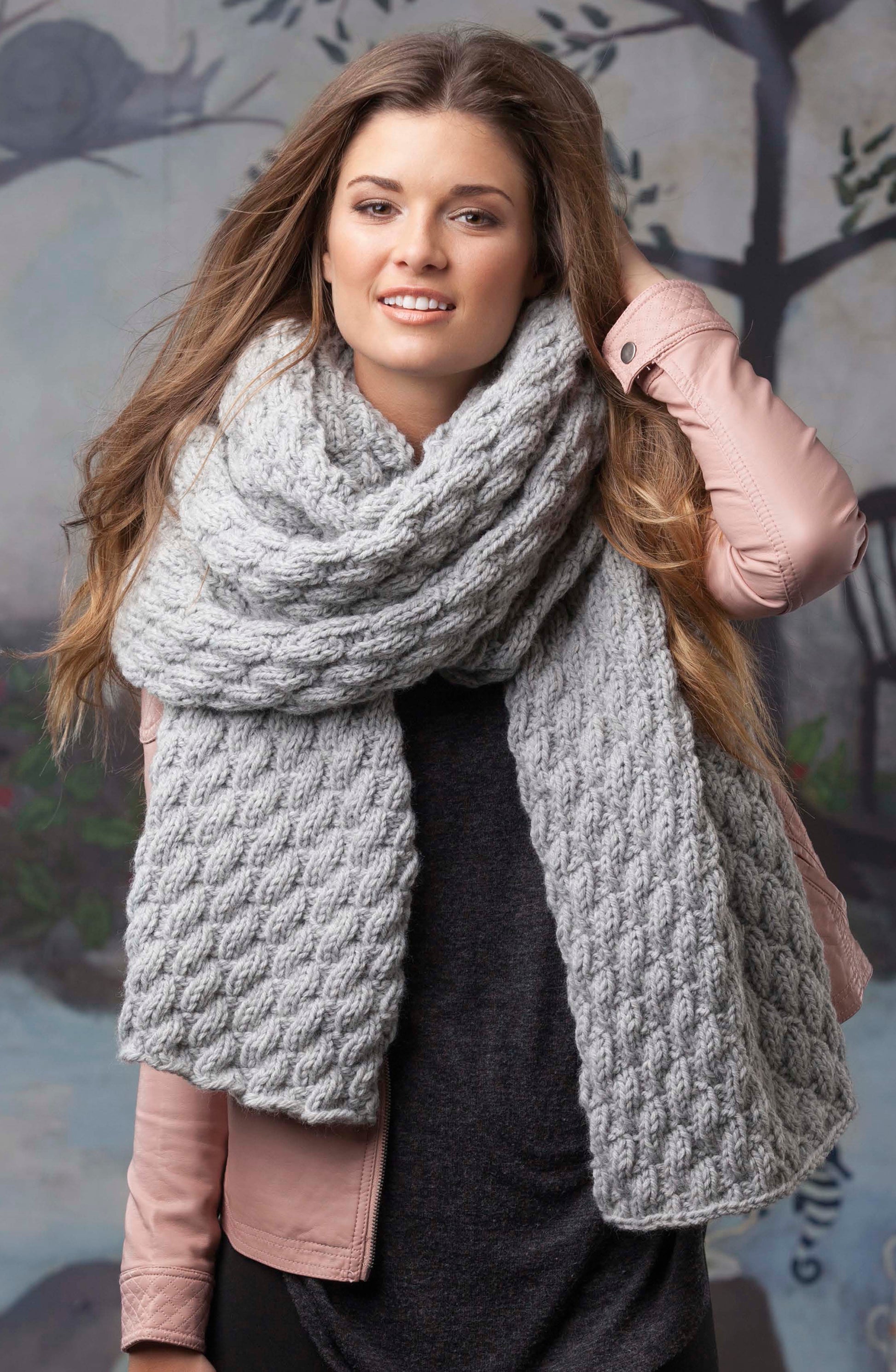 A woman with long brown hair smiles while wearing a chunky light gray scarf from Super Scarves to Knit by Leisure Arts, paired with a pink jacket and dark top. She holds her hair, with trees and painted scenery blurred in the background.