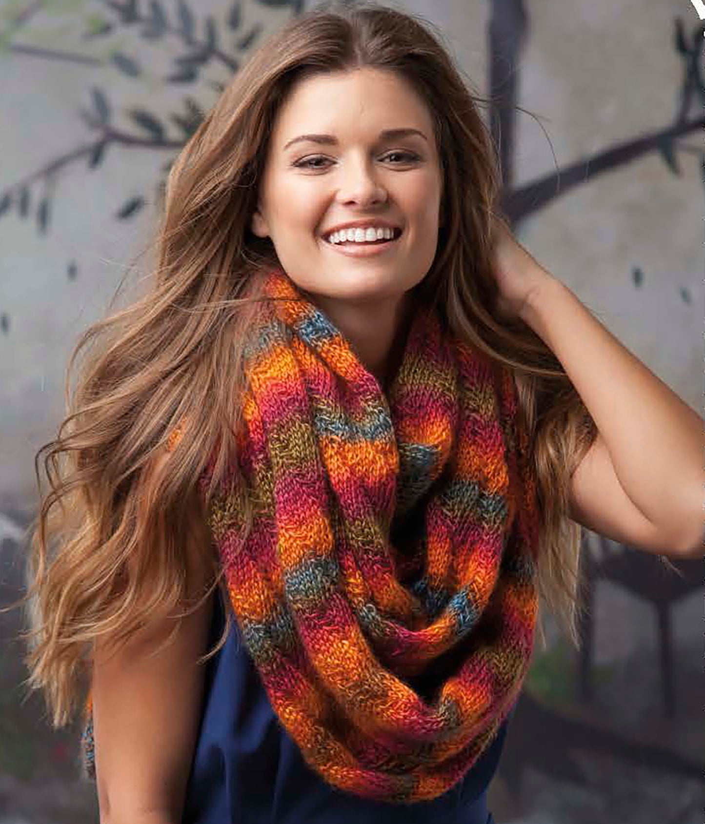 A smiling woman with long brown hair wears a vibrant, colorful scarf from Leisure Arts' "Super Scarves to Knit," featuring intricate patterns, as she stands outdoors against a softly blurred background.