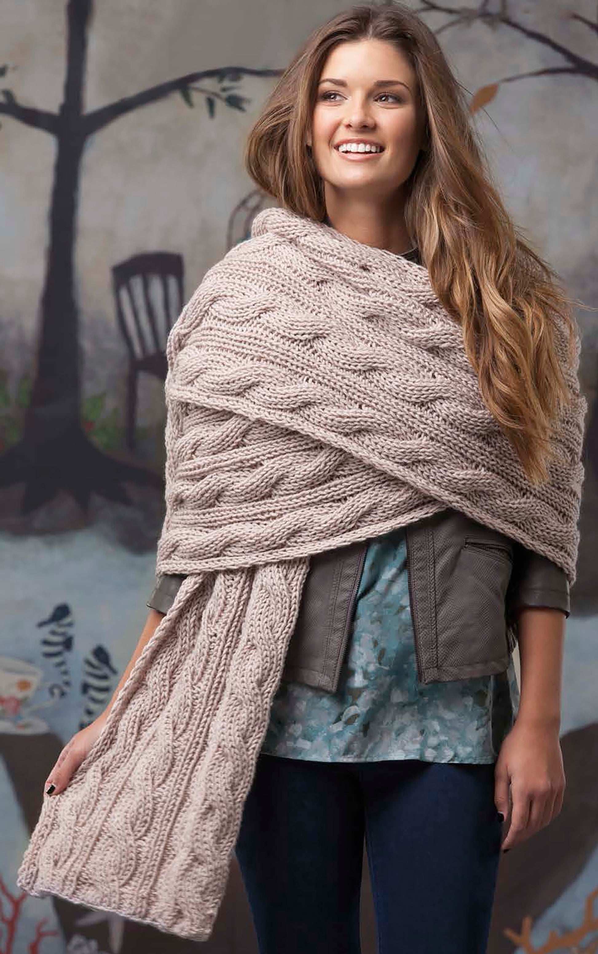 A woman with long hair smiles while wearing a chunky knit beige scarf made from bulky yarn, created with Super Scarves to Knit by Leisure Arts, over a gray jacket and blue top, standing in front of a mural with trees and chairs.