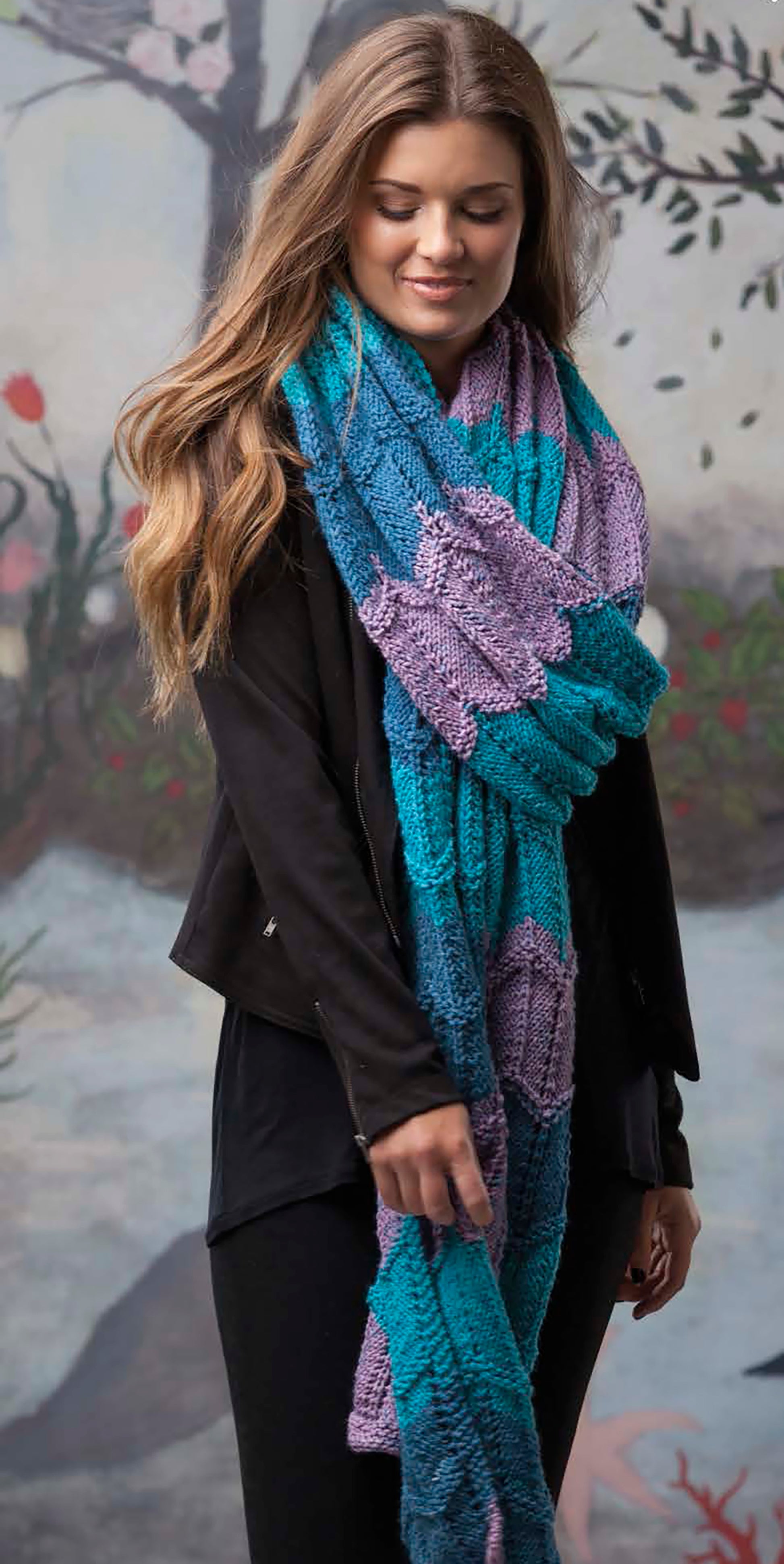 A woman with long, wavy brown hair wears a black outfit and a blue-purple bulky scarf from Leisure Arts' "Super Scarves to Knit," standing before a painted outdoor backdrop.