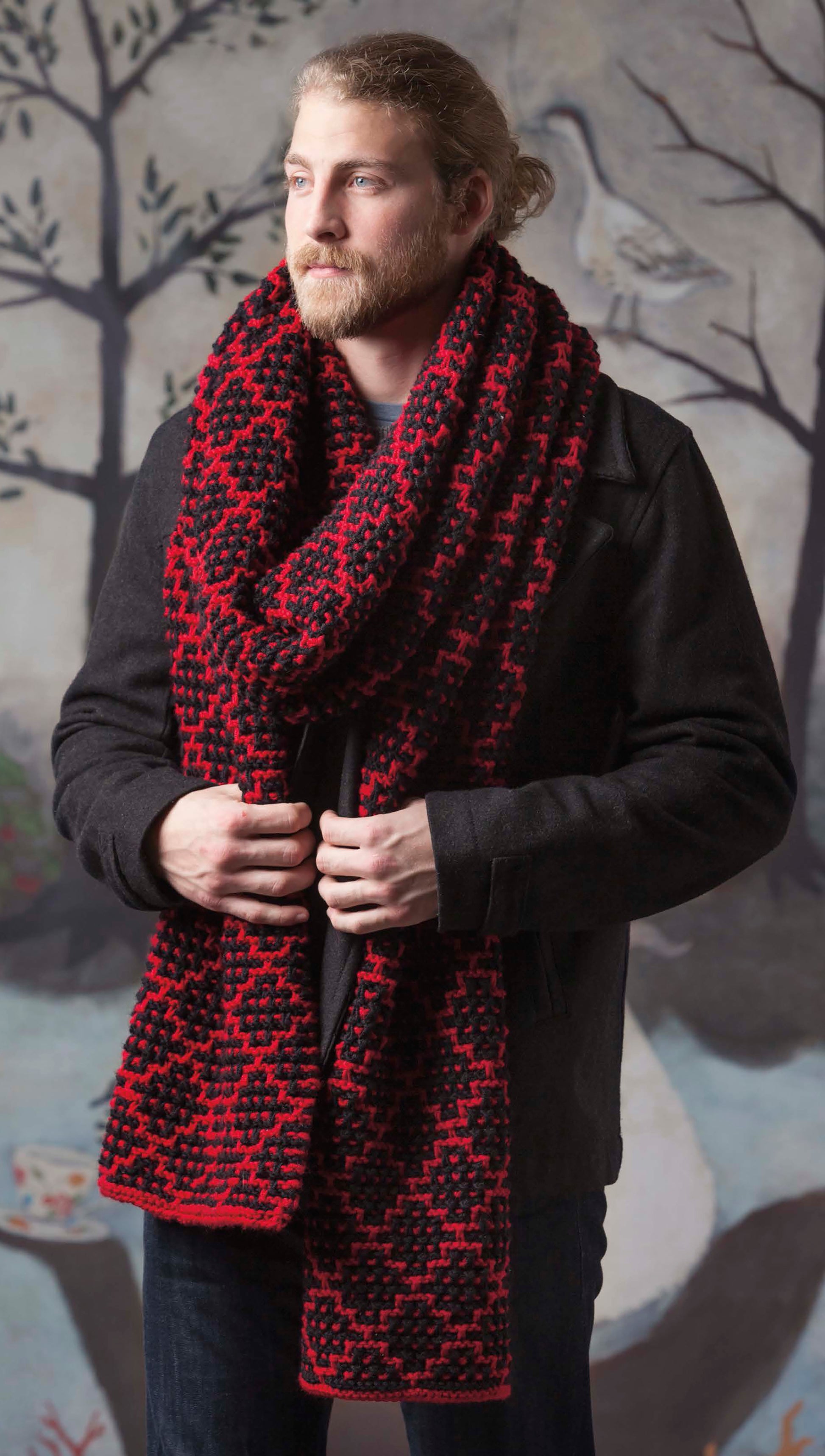 A man with a beard and tied-back hair models a bold, red and black scarf with striking knitting patterns from Leisure Arts' "Super Scarves to Knit," worn over a dark jacket, standing before a backdrop of painted trees and birds.