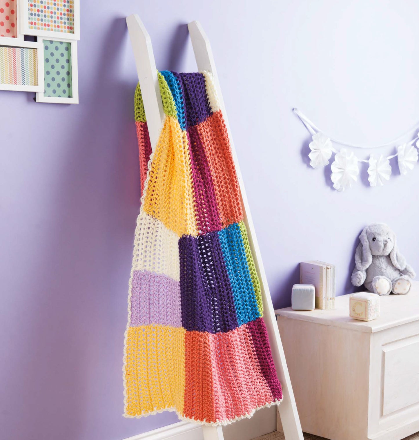 The Leisure Arts Color-Block Baby Blankets, crafted from medium weight yarn, hang over a white ladder in a pastel nursery with a stuffed bunny, books, and decorative accents—perfect for adding cozy style to your home décor.