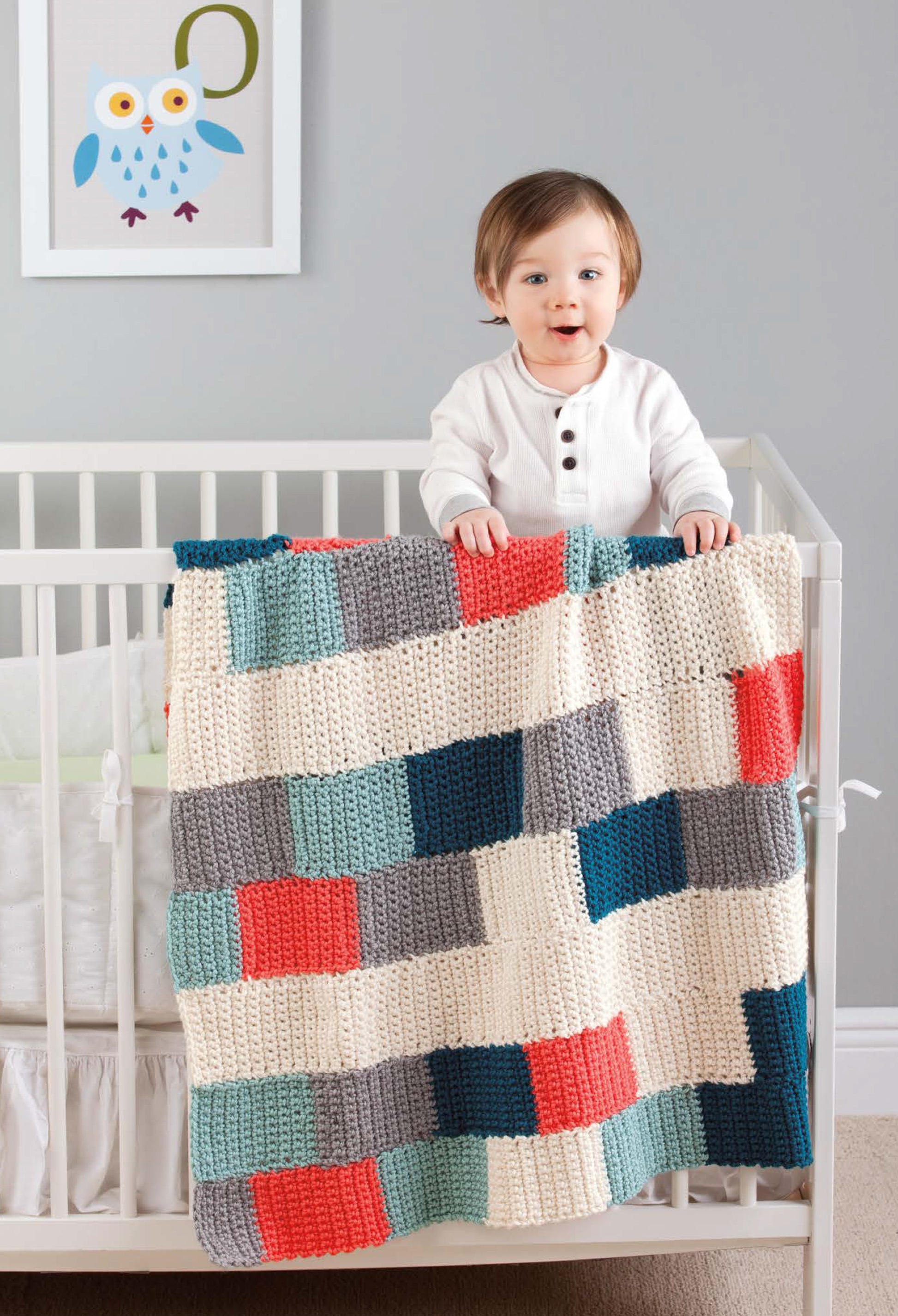 A young child in white pajamas stands in a crib, holding a Leisure Arts Color-Block Baby Blanket. The colorful patchwork design adds charm to the nursery, complementing the home decor and owl wall art.
