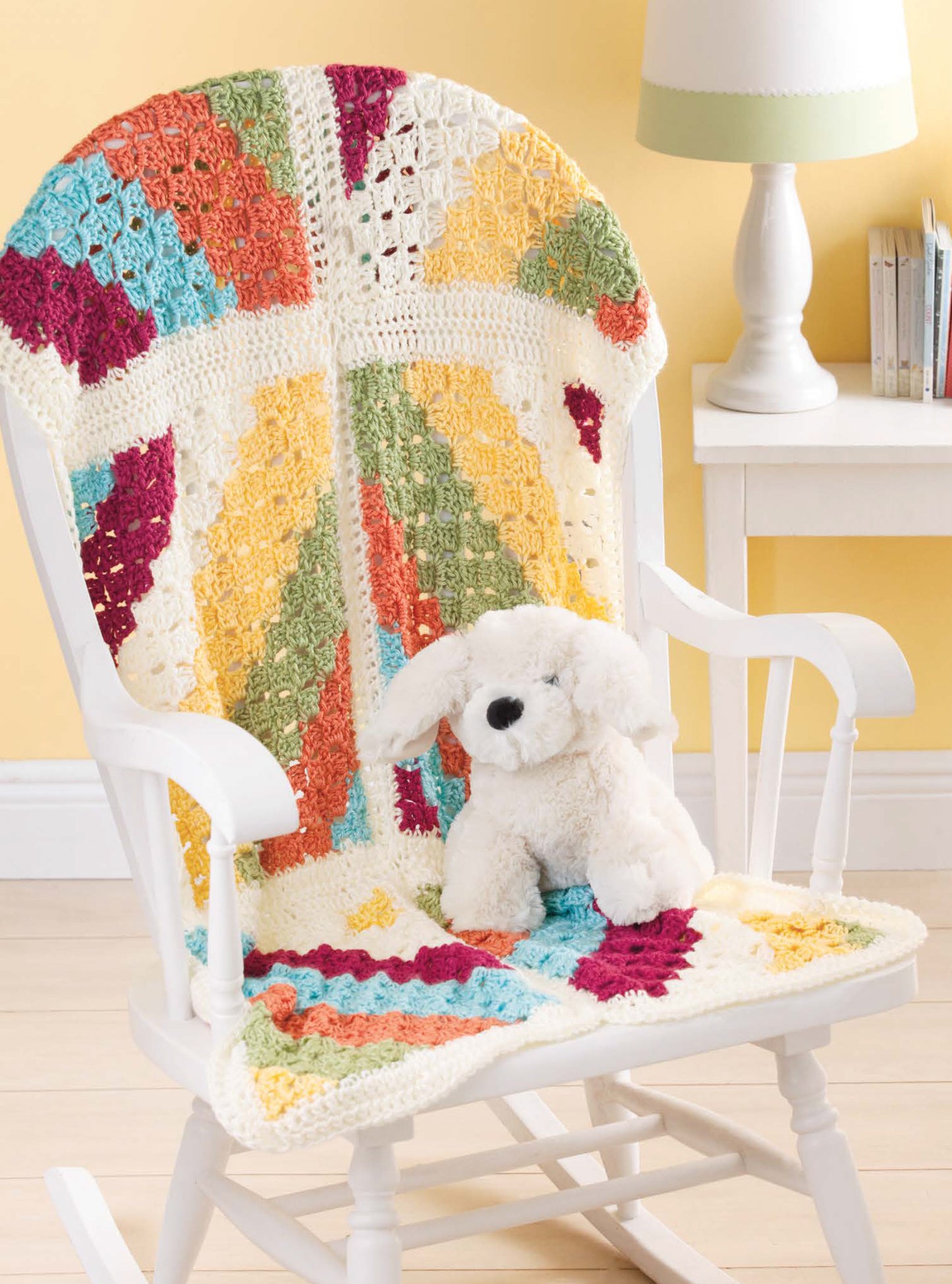The Color-Block Baby Blankets by Leisure Arts, crafted from medium weight yarn, add a cheerful touch draped over a white rocking chair with a plush dog toy, complemented by a white table and lamp against a sunny yellow wall.
