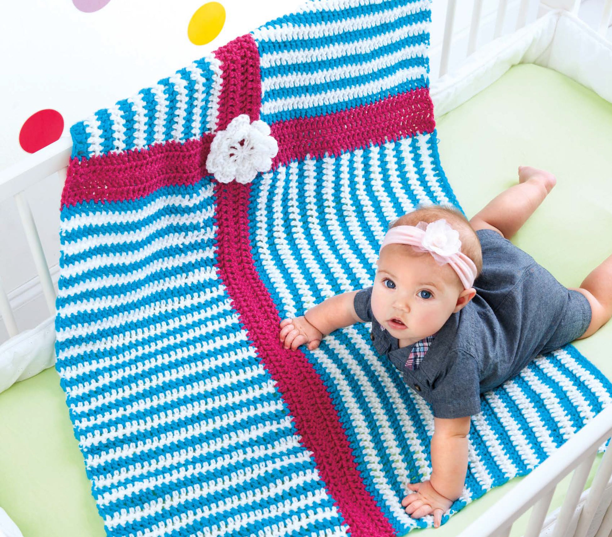A baby in a grey outfit and pink headband lies on a Leisure Arts Color-Block Baby Blanket, a striped blue, white, and pink crochet design with a white flower—charming decor for any crib.